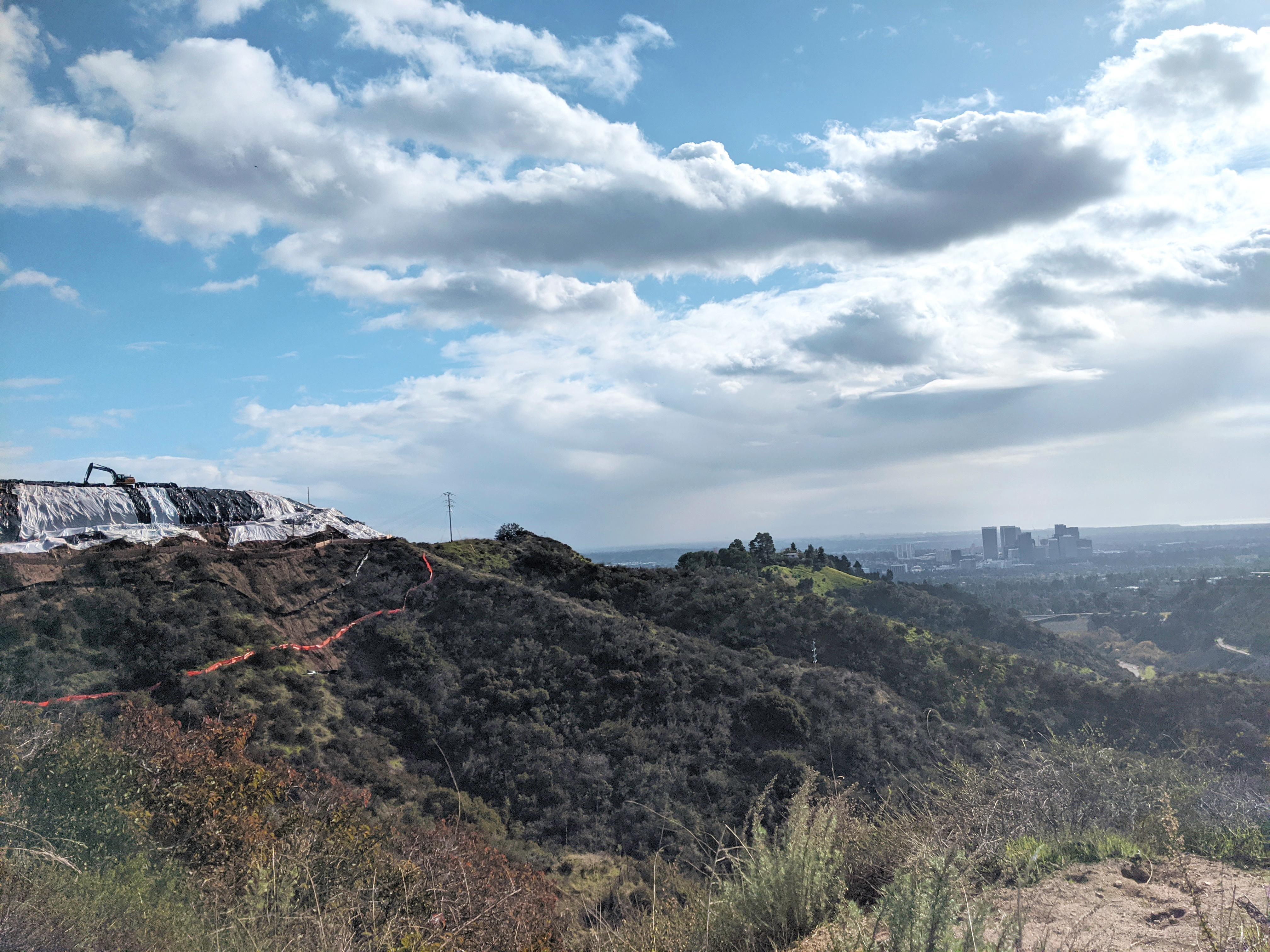 A hillside with a bunch of plastic on it and an earthmover atop it. In the backround, there is a view of the city, partly obscured by smog, and the outlines of a cluster of tall buildings.
