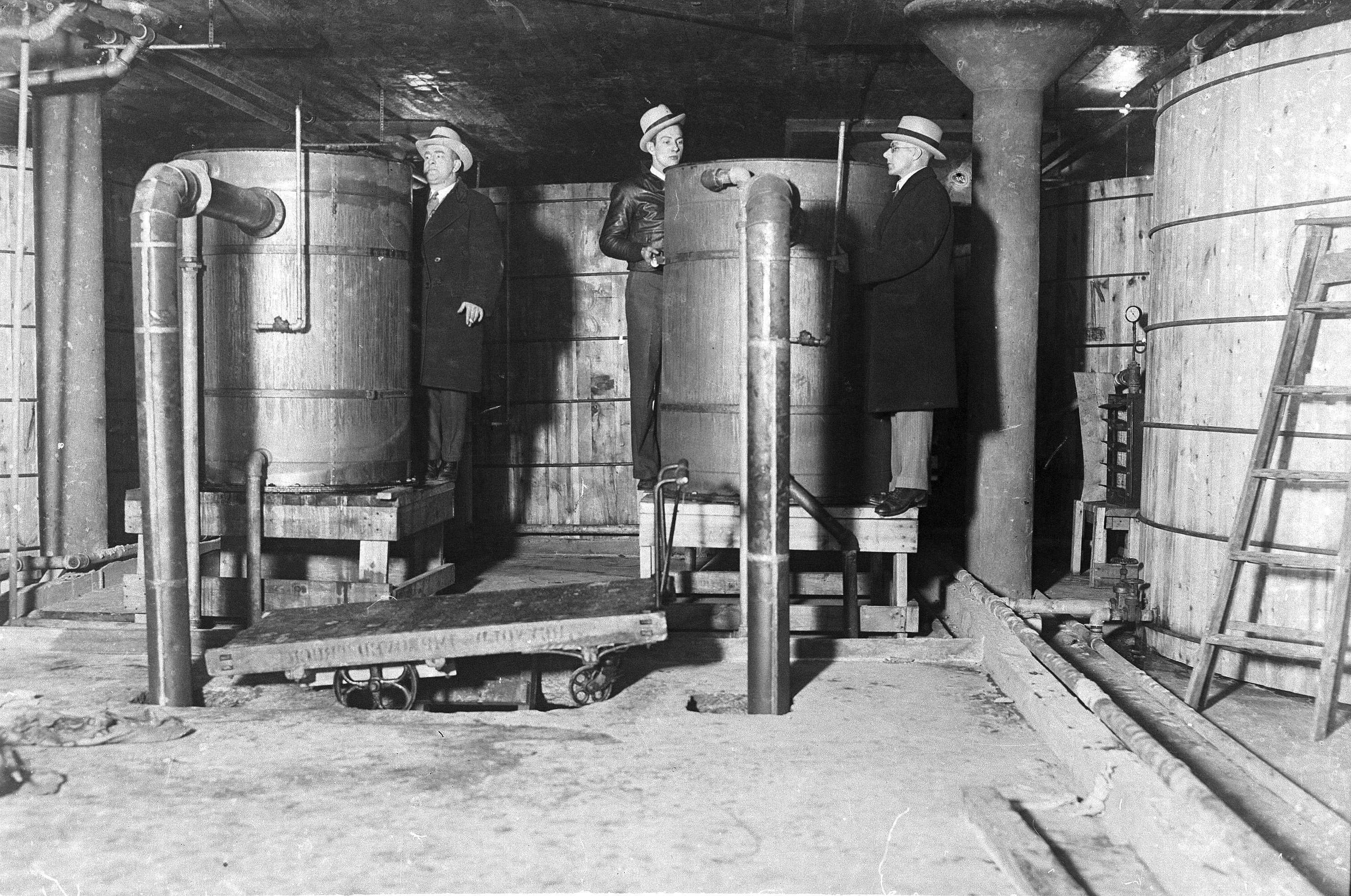 A black and white photo of three men in trench coats and fedoras next to big barrels.