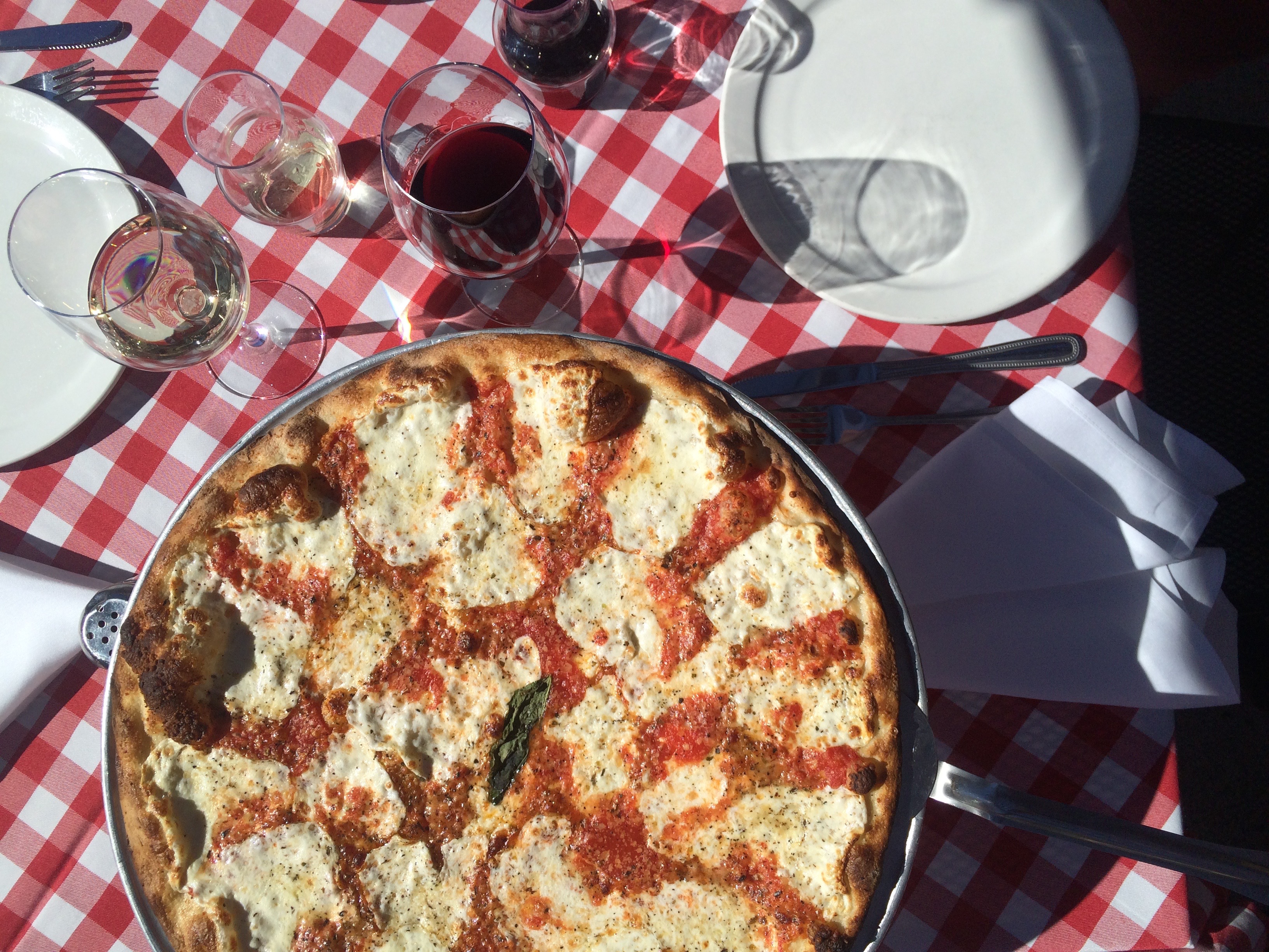 A pizza with mozzarella and red sauce sits on top of a red-checkered tablecloth, with glasses of wine.