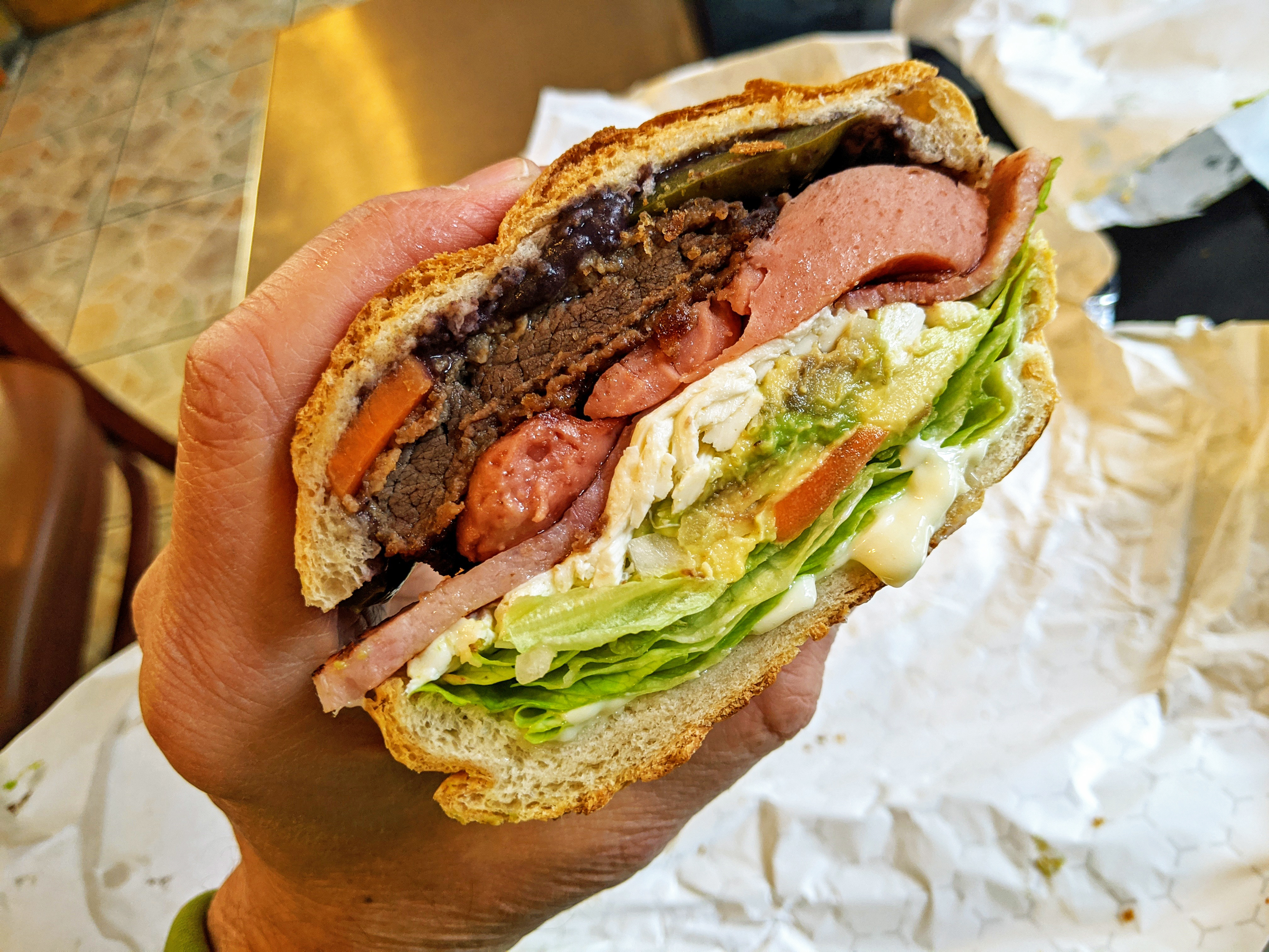 A hand holds a bulging sandwich with many colorful layers.