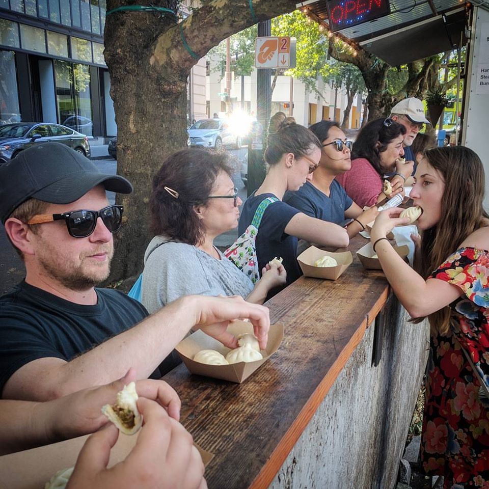 A line of customers eat bao over a wooden railing at the original cart