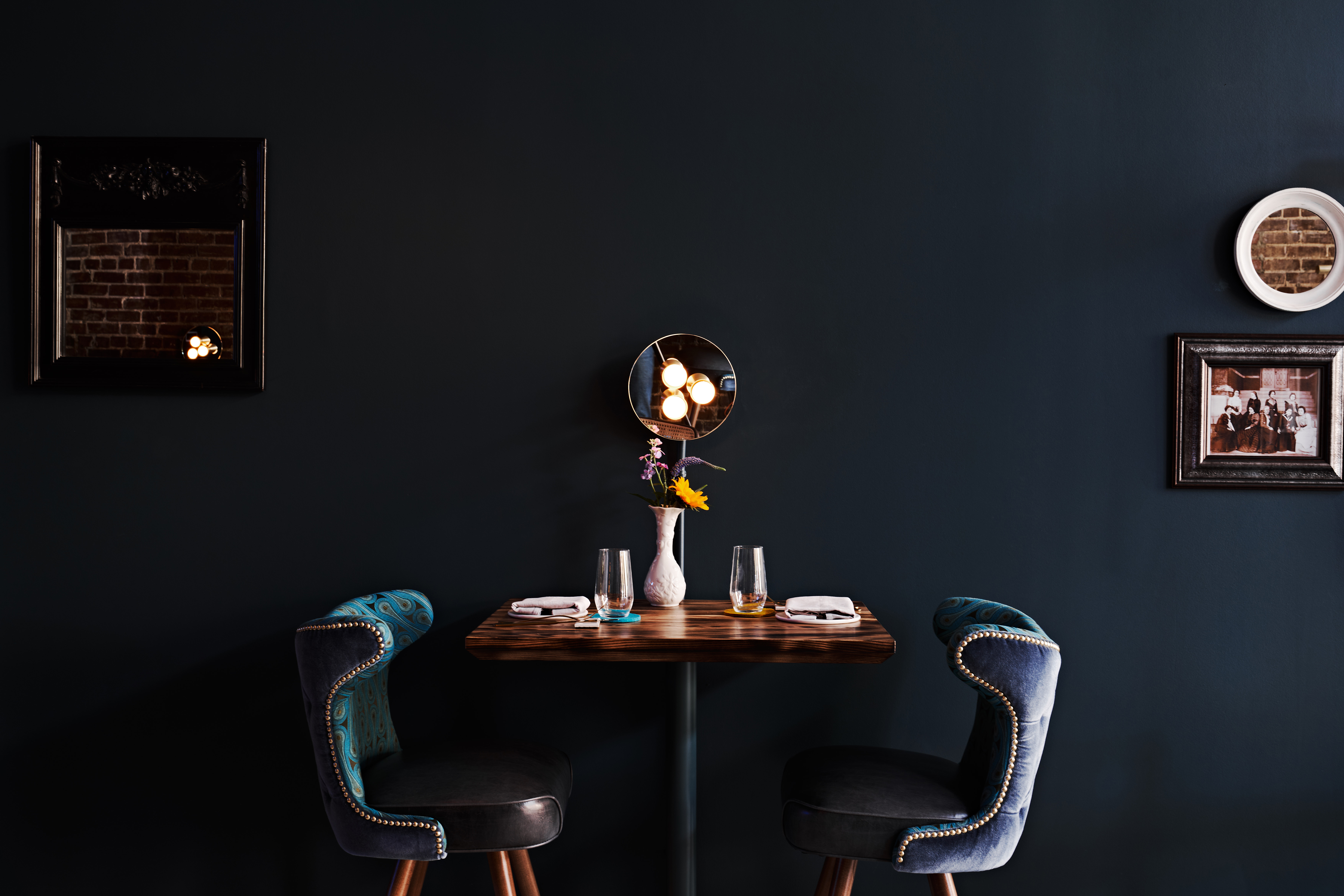 a wide shot of a cocktail round with two blue velvet tufted chairs against a dark navy wall