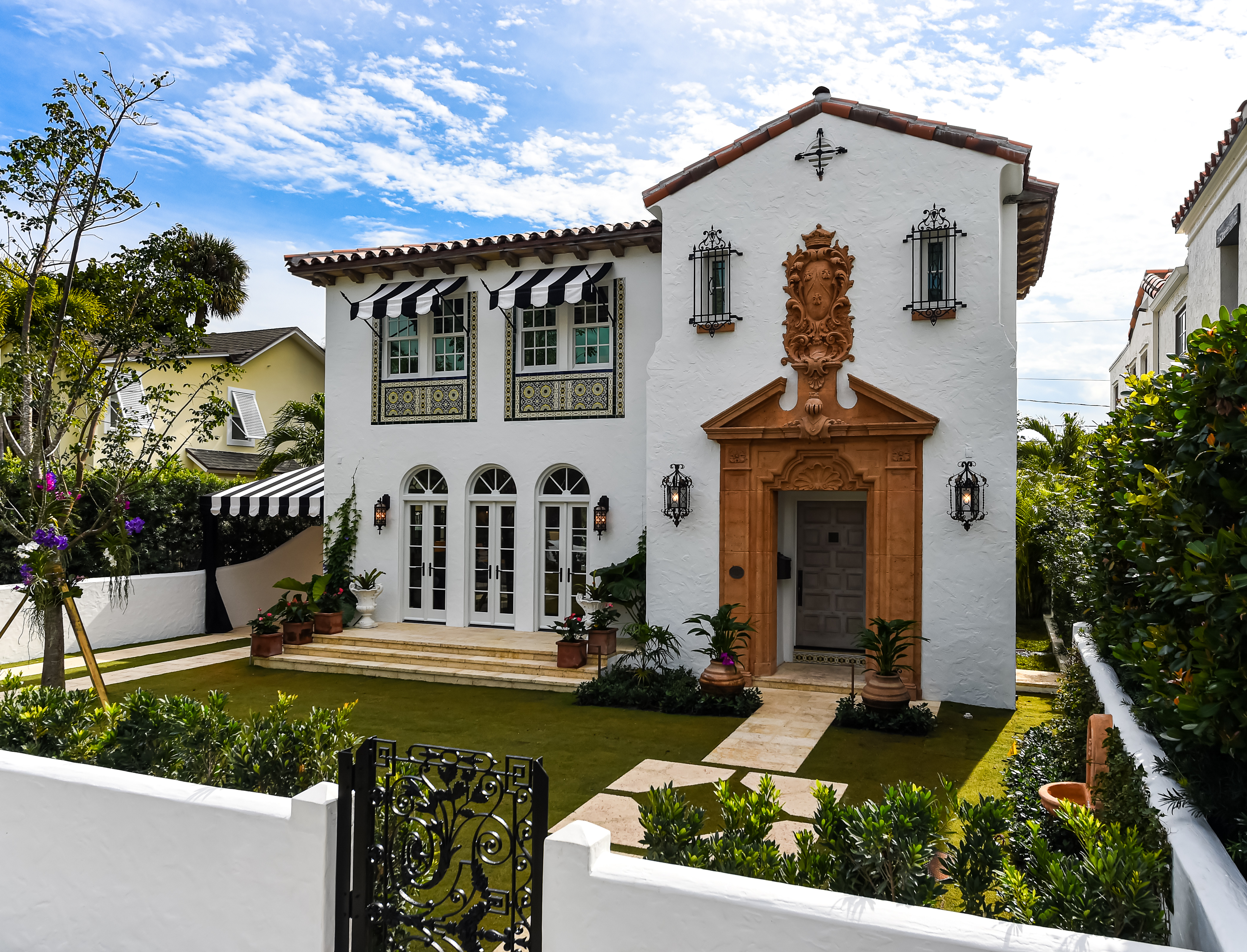 A Spanish style home features white stucco, striped awnings, and an elaborate wood portico at the entrance. 
