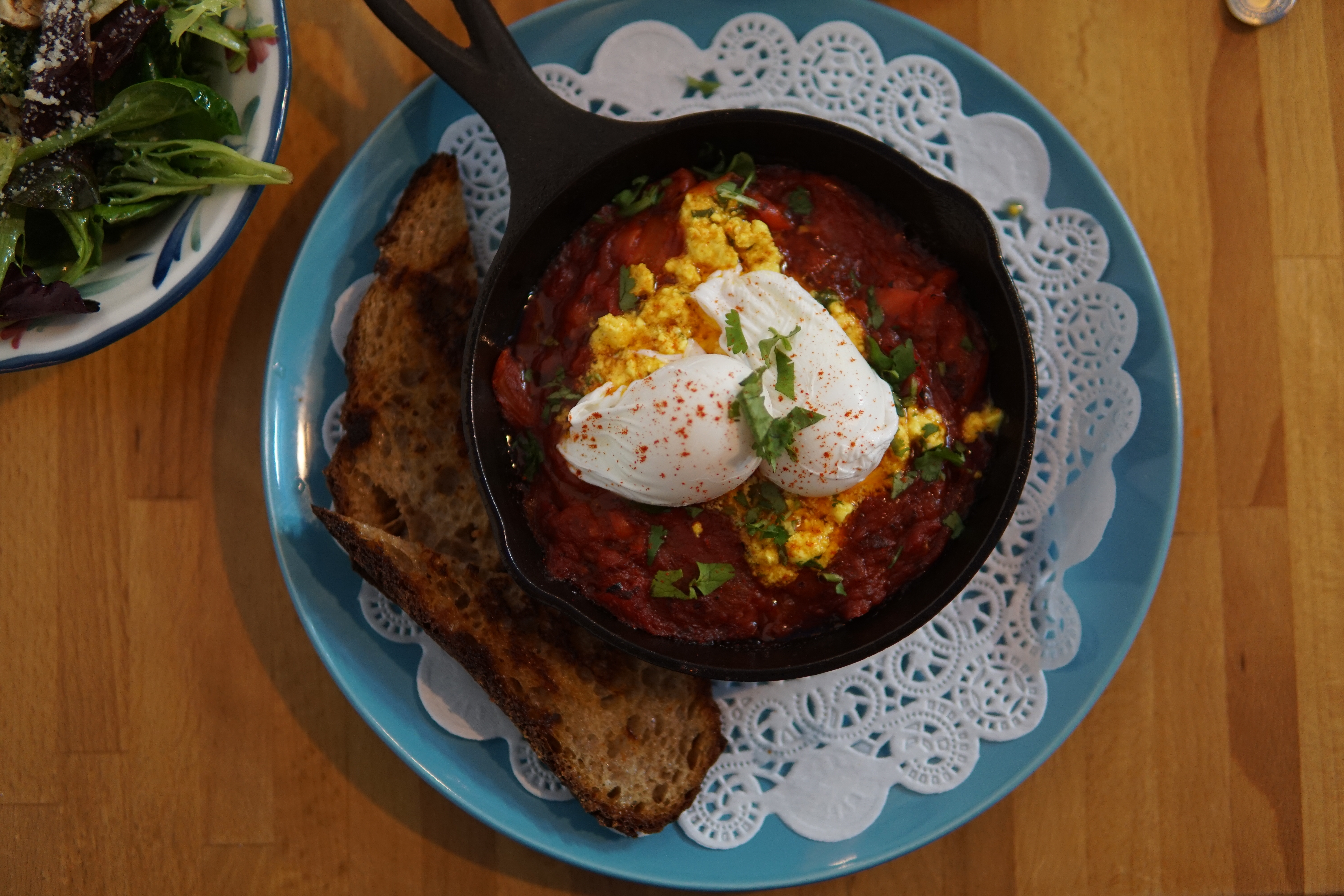 Two poached eggs sit in a cast-iron skillet, which sits on a blue plate with a doily and two pieces of toast