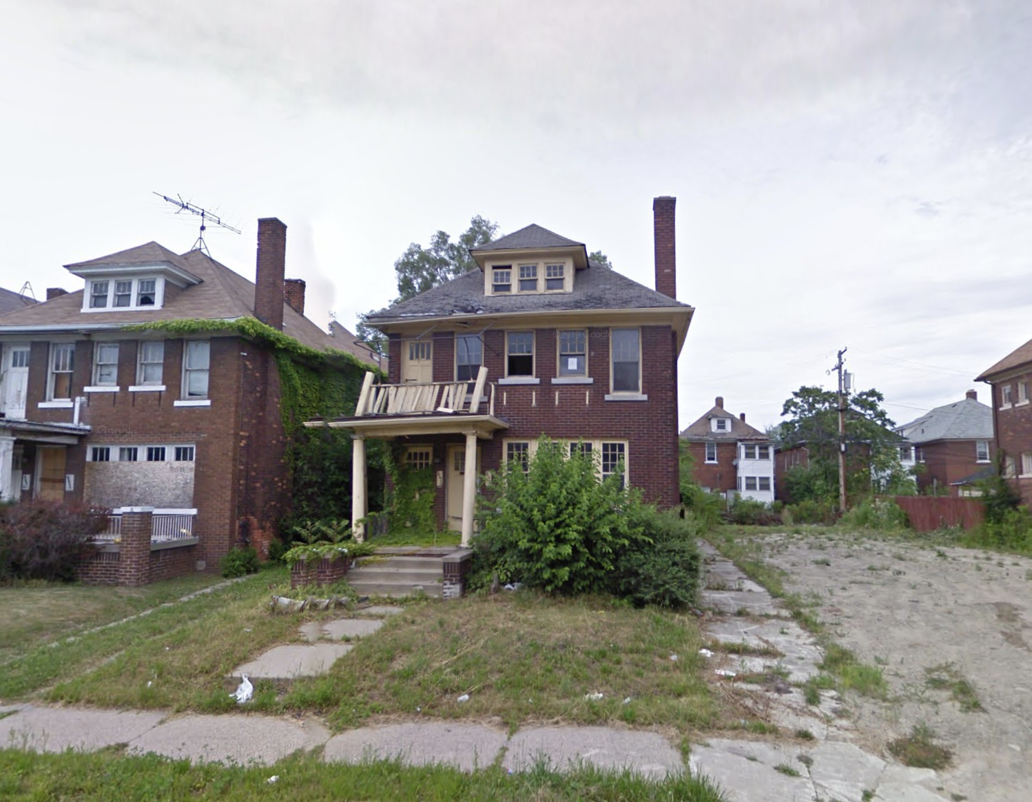 A two story brick home. There’s a large untended bush outside the front windows and a wobbly porch overhang. 