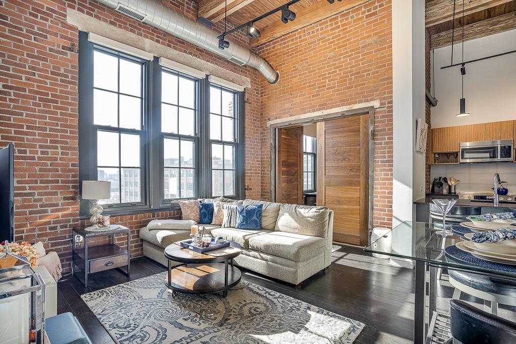 A living room with very high ceilings and very big windows, with furniture and exposed beams and ducts. 