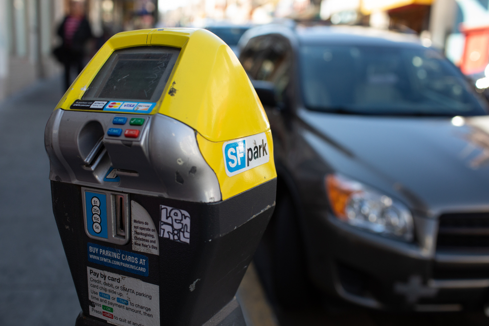 A closeup of a yellow parking meter with a blue and white SF Park sticker on the side, parked near a black car at the curb.