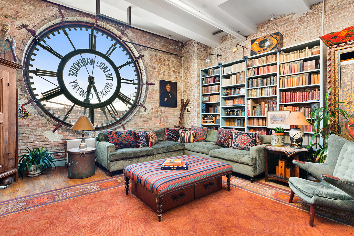A living area with exposed brick, a large clock with views of the Brooklyn Bridge, a green couch, blue bookshelves, and beamed ceilings.