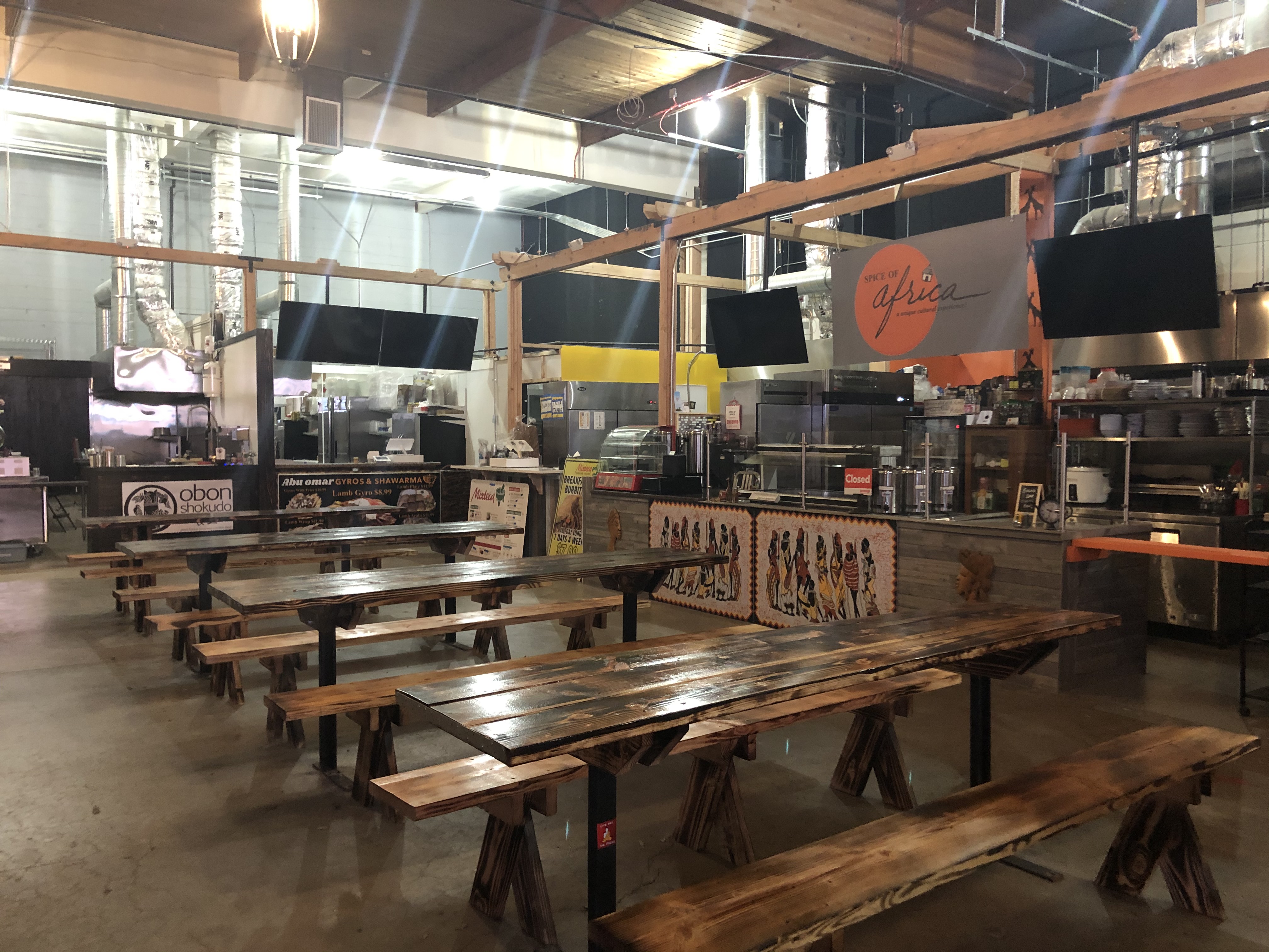 Picnic tables sit in the middle of an industrial, warehouse-like market. Stalls sit around the chairs and tables.