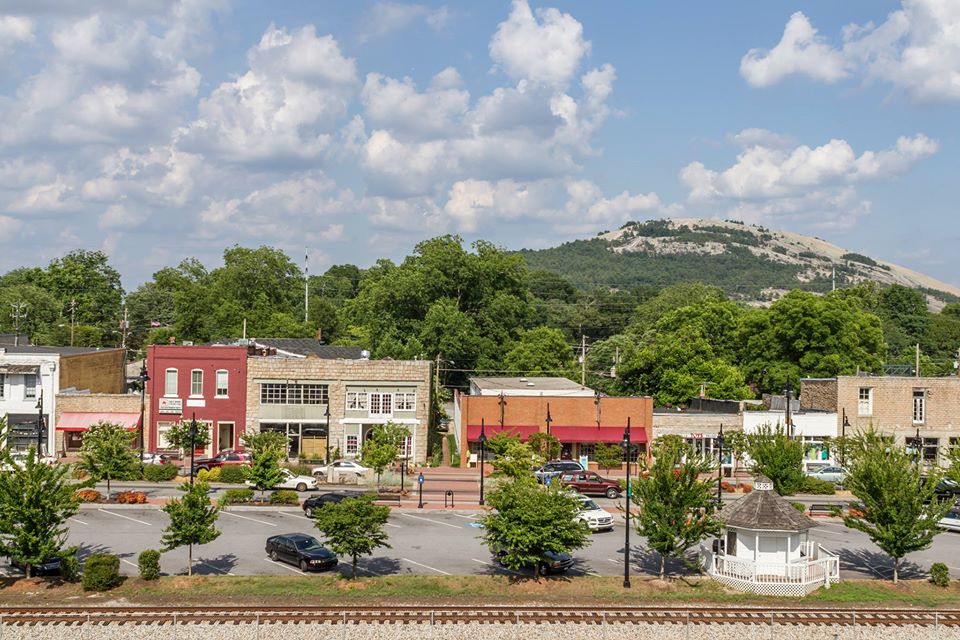 A row of old storefront between a parking lot with a few cars and a large granite mound, with a blue sky overhead. 