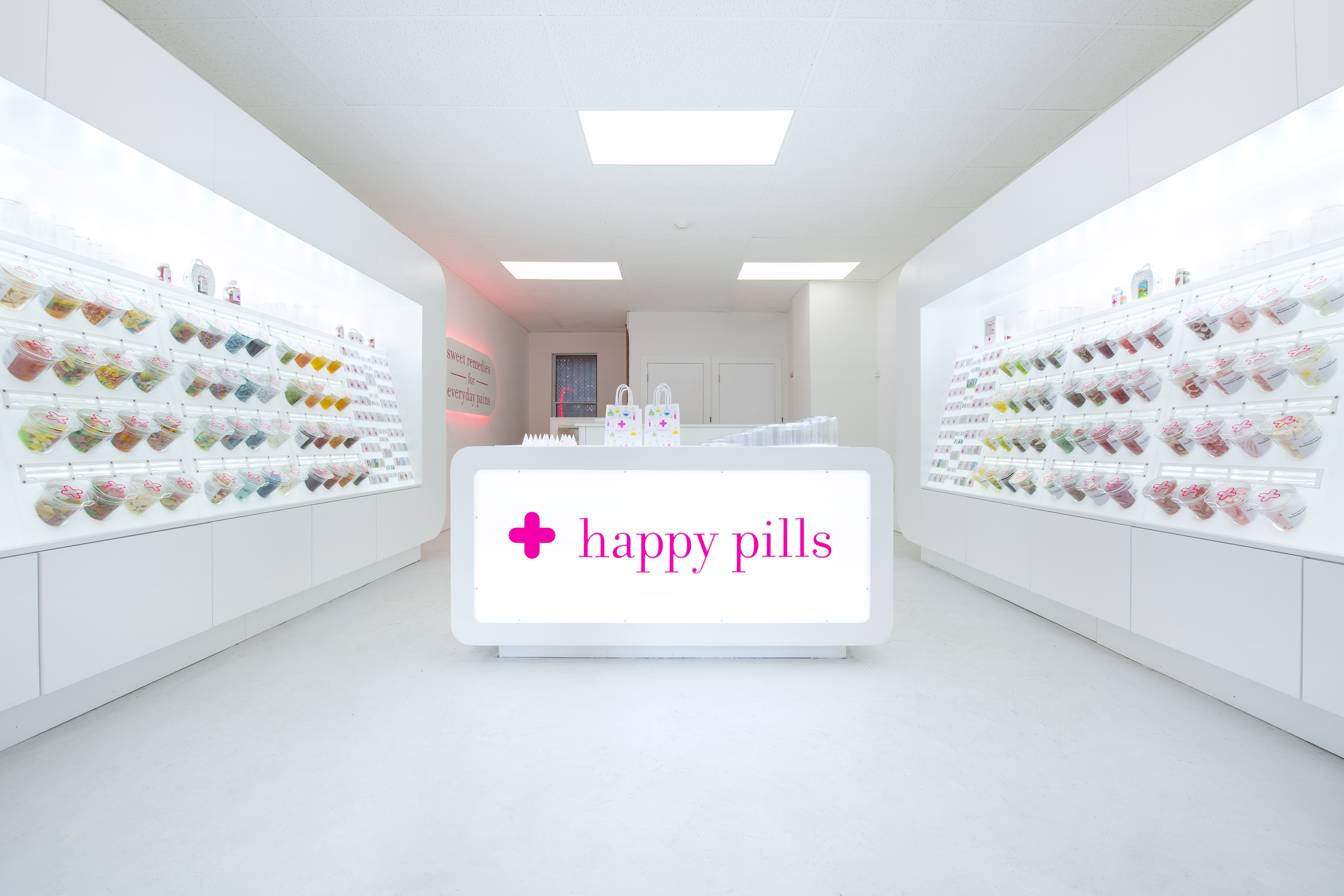 Interior of a bright white candy store with a pharmacy theme. A counter in the middle has a pink logo that reads “Happy Pills,” and each wall is lined with bulk candy containers.