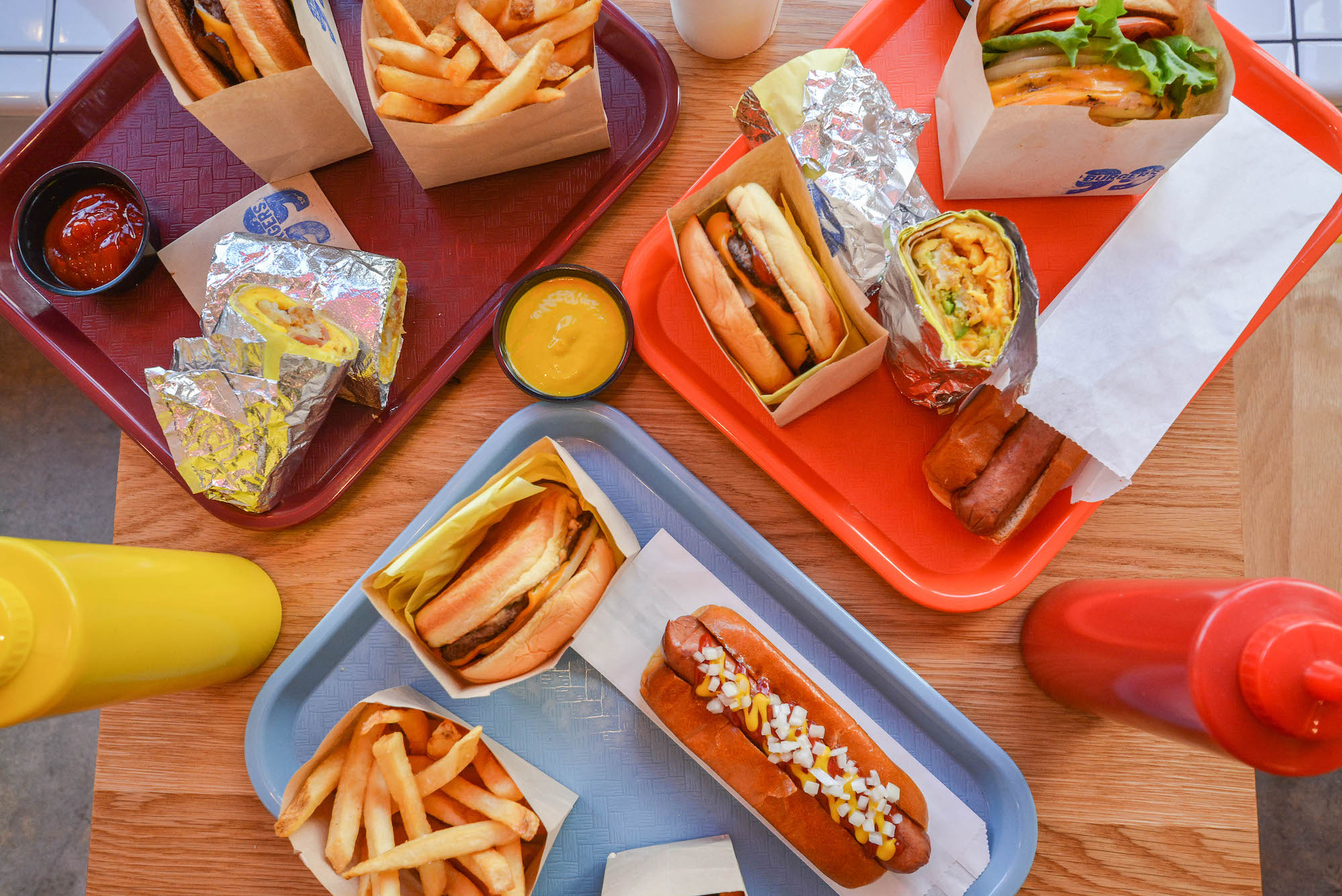 An above shot of three colored trays of burgers and hot dogs and fries.