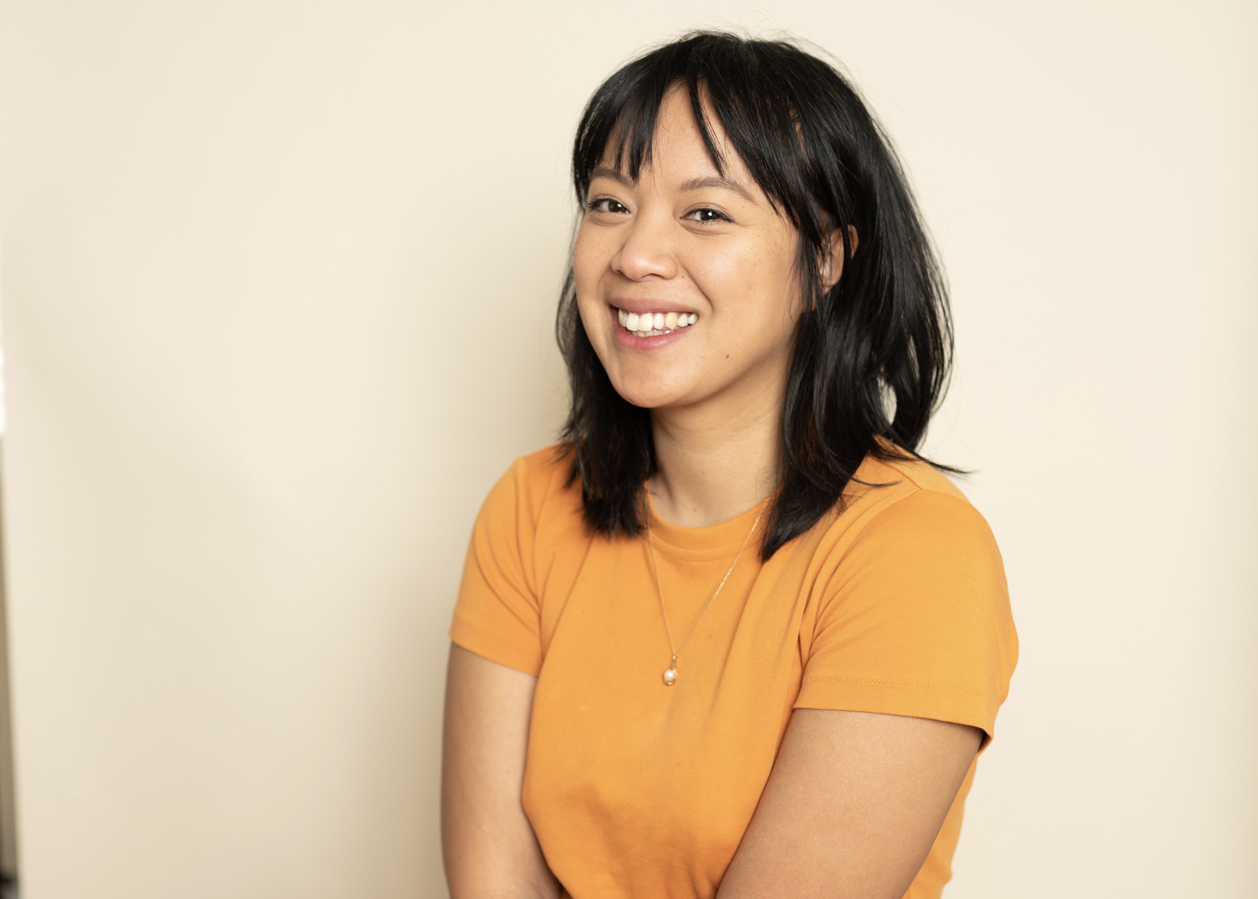 Diane Lam sits in front of a beige wall wearing a yellow-orange shirt. She is a young Cambodian-American woman with black hair and bangs.