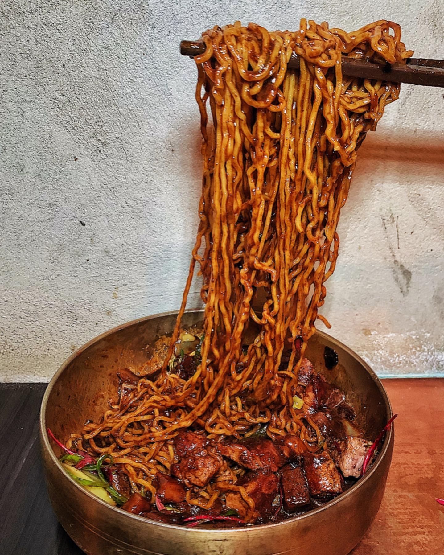 A metal bowl with noodles and steak inside, with noodles being pulled up.