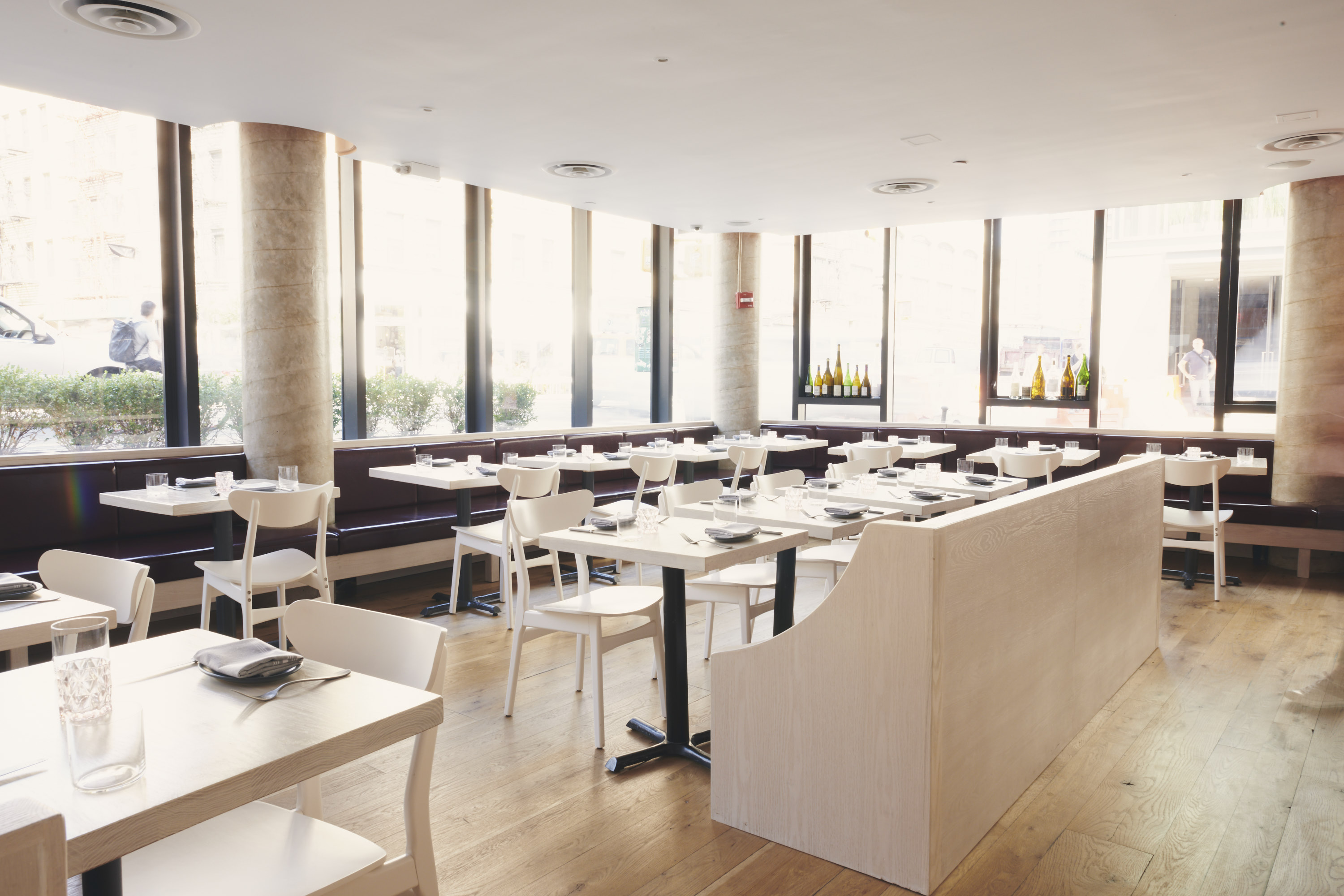 The interior of a restaurant with floor to ceiling windows, off white tables and tables and chairs, and a low ceiling