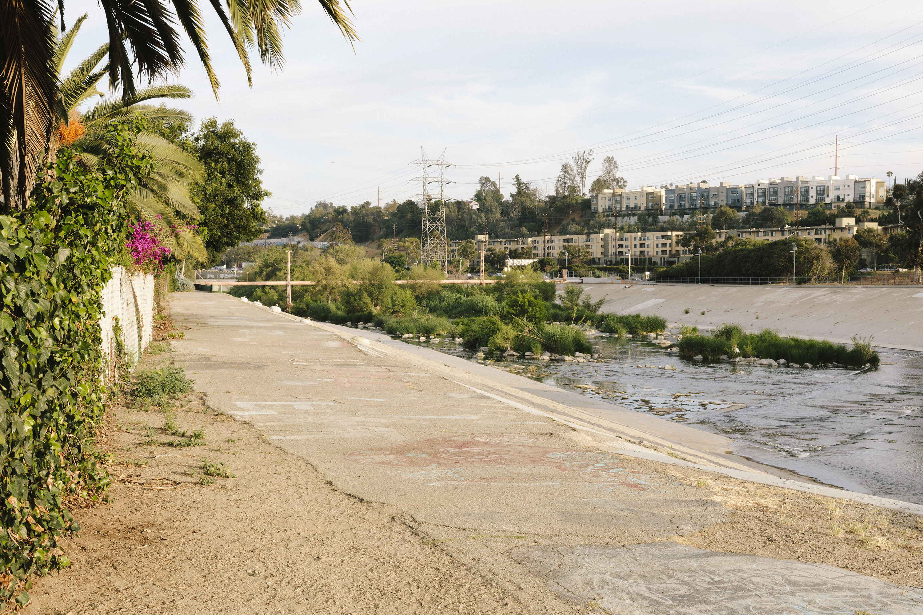 A sidewalk path along a riverbed. There are buildings in the distance.