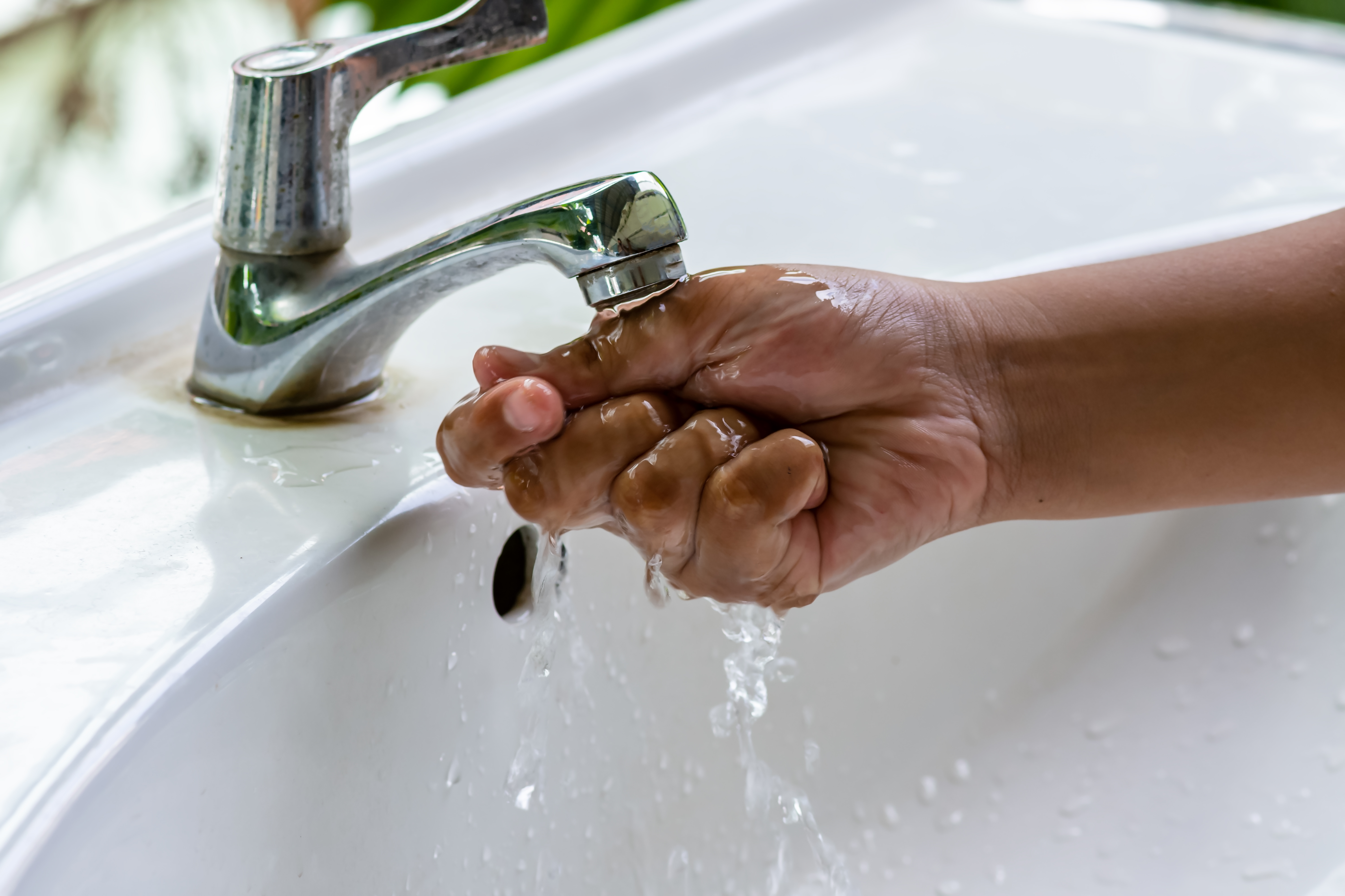 A hand with short nails runs under a water faucet in a white sink.