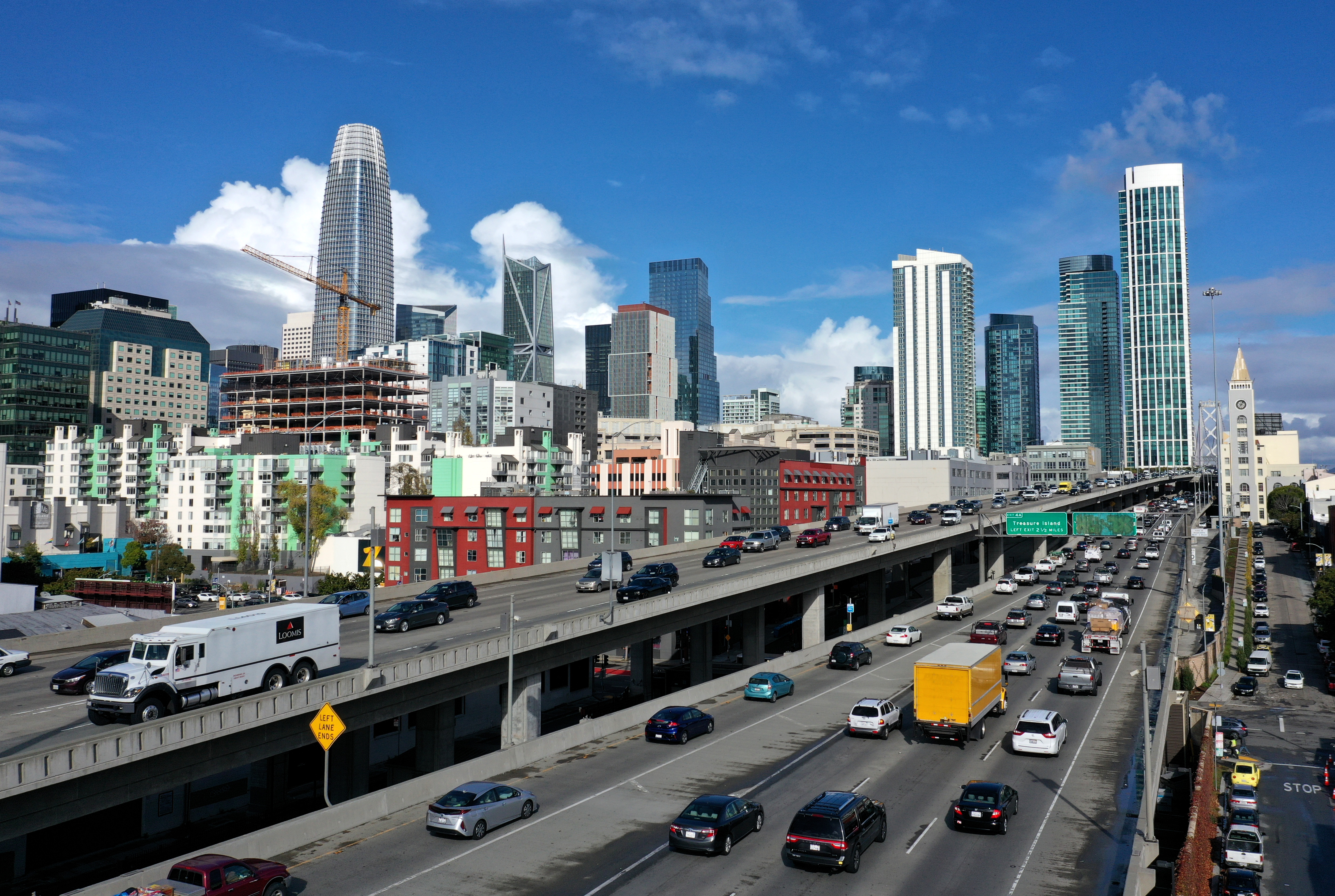 Traffic on a busy freeway, photographed from above, with tall, glass-encased high-rises visible on the horizon.
