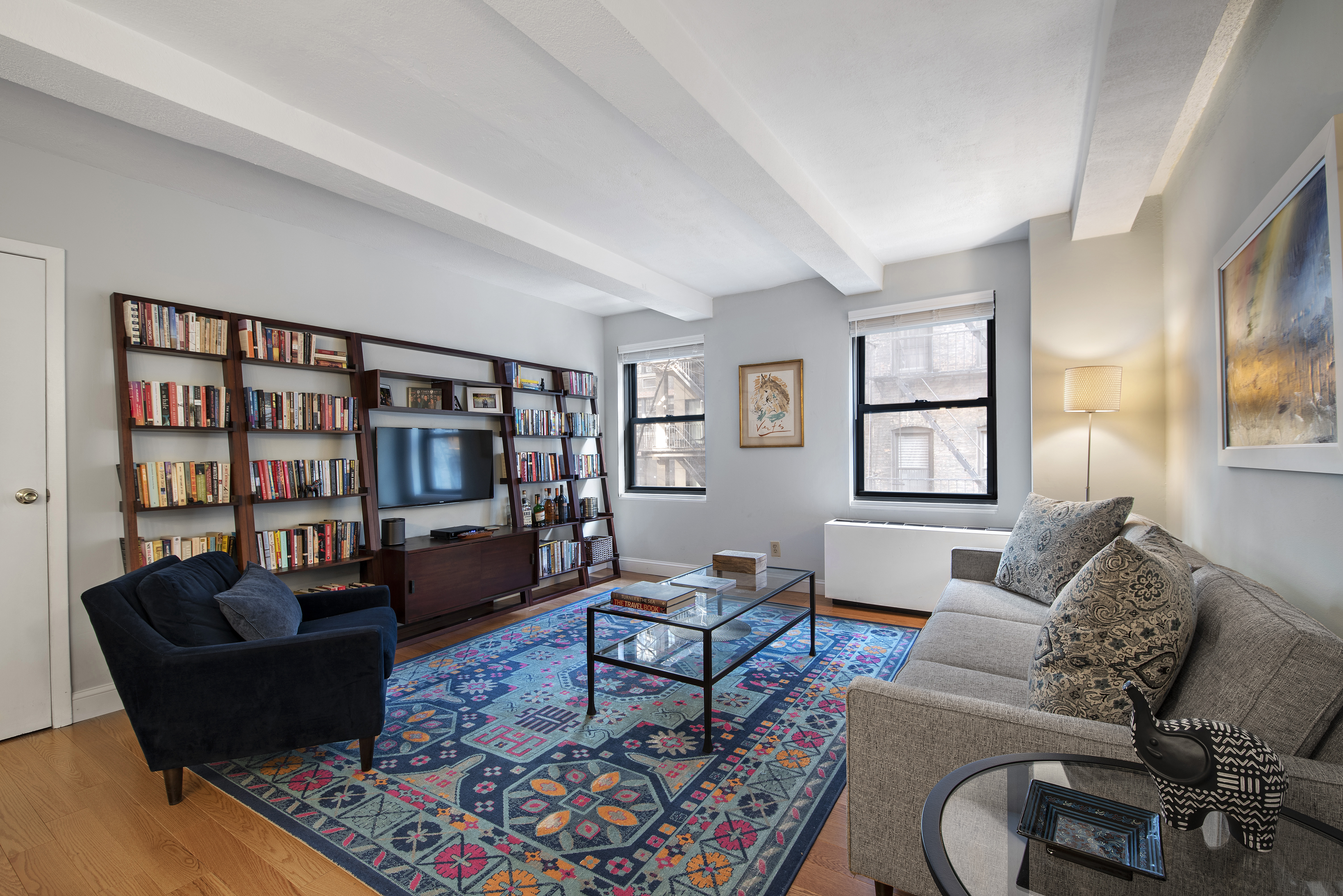 A living area with hardwood floors, beamed ceilings, several bookshelves, and a colorful rug.