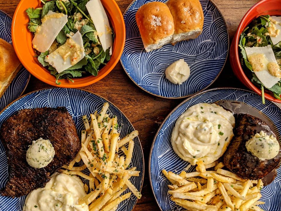 Overhead view of steak, mashed potatoes, fries, rolls, and salad on blue and orange plates on a wooden table