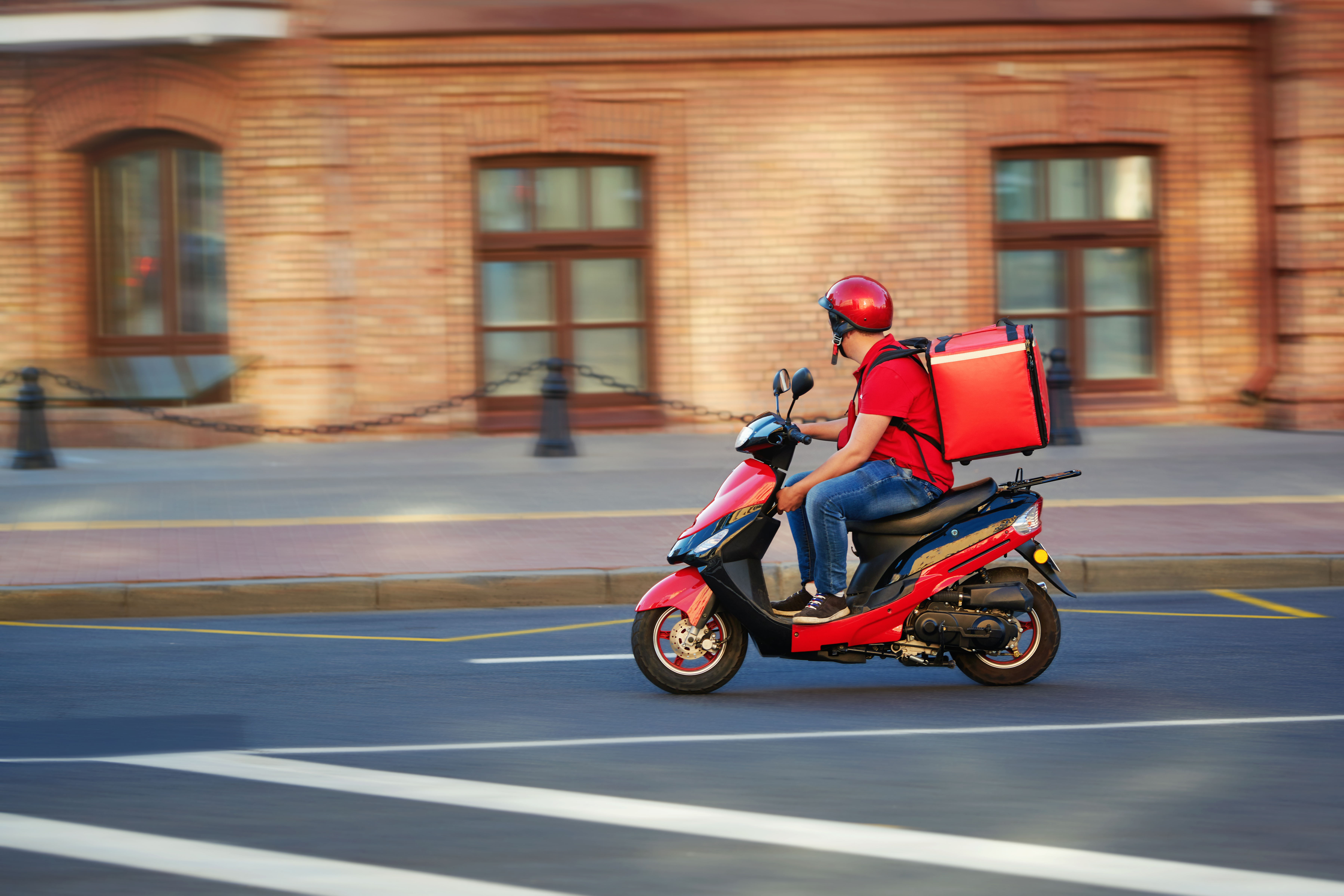 A biker in a red outfit bikes down an empty street with a red food delivery bag on his back