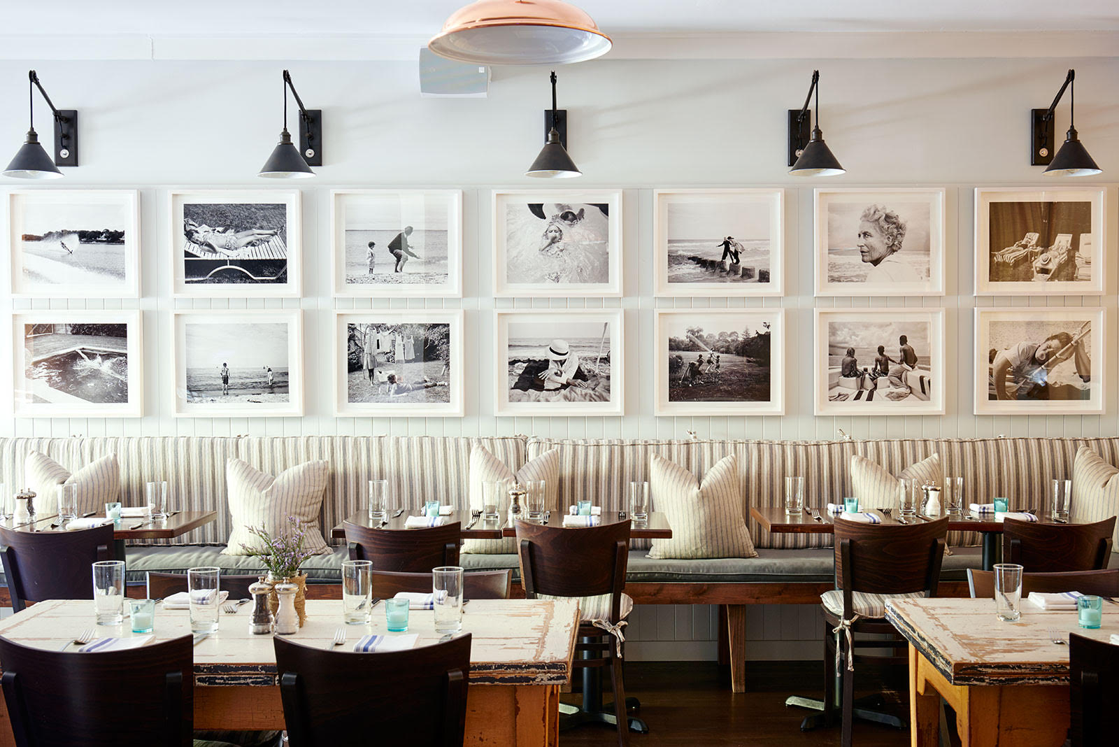 A white dining room with a wall of white framed photos over a banquette with pillows.