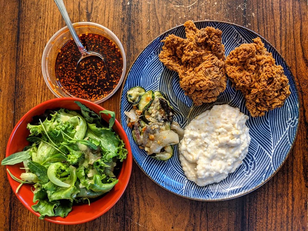 Overhead view of fried chicken, grits, and roasted zucchini on a blue plate. A red bowl of greens sits to the side, along with a plastic container of a red sauce. All sit on a wooden tabletop.