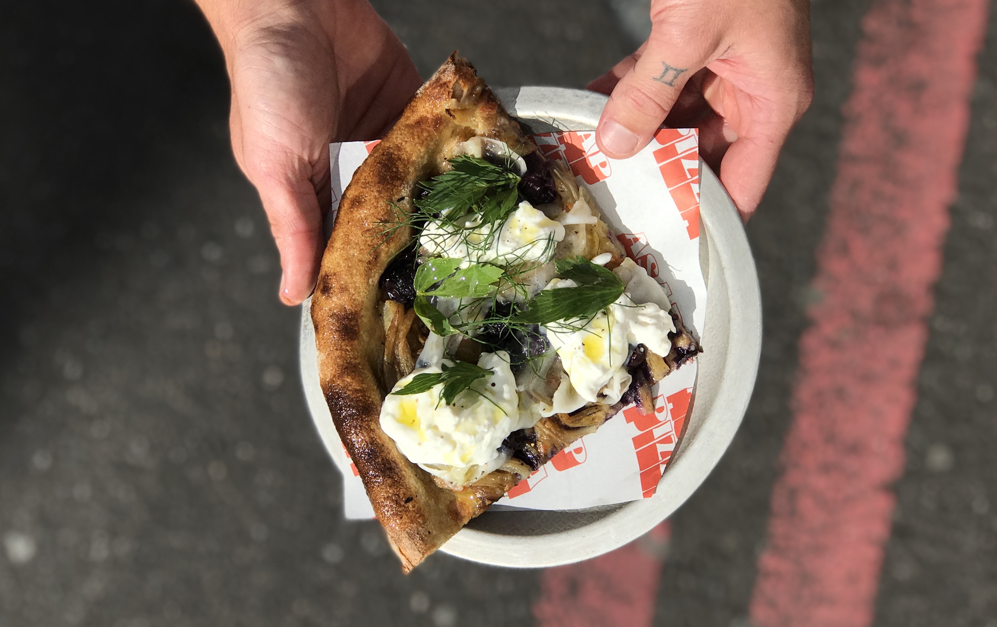 ASAP Pizza Borough Market shows off a slice of pizza with fragola grape, stracciatella, and fennel fronds held from a birdseye view over a road