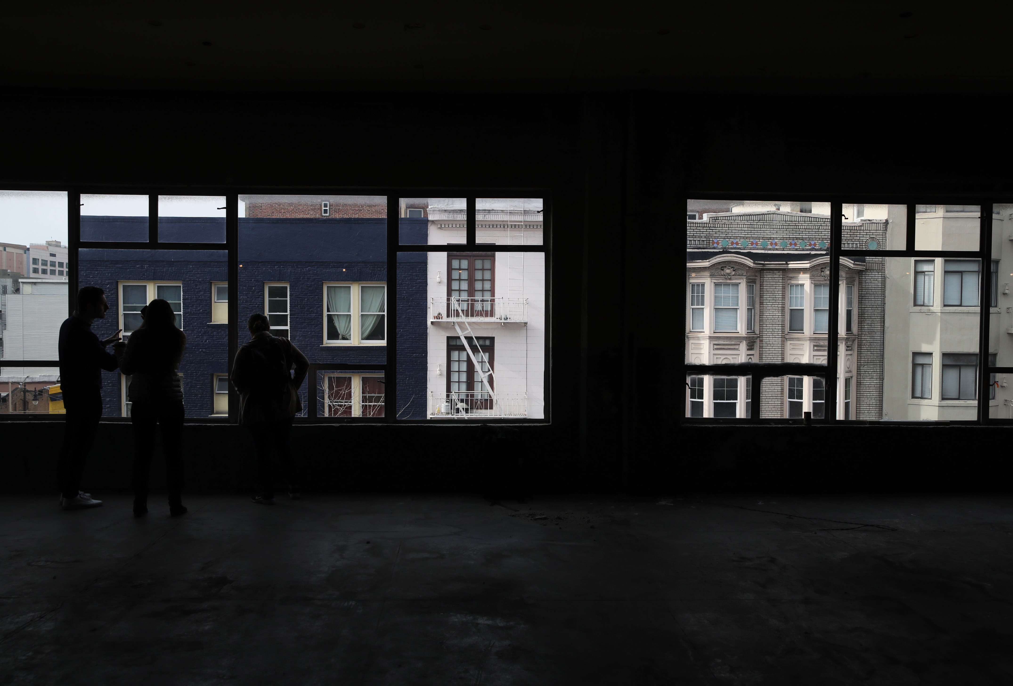 Four silhouettes of people standing in the middle of an almost entirely empty room, looking out a row of windows that make up one entire wall.