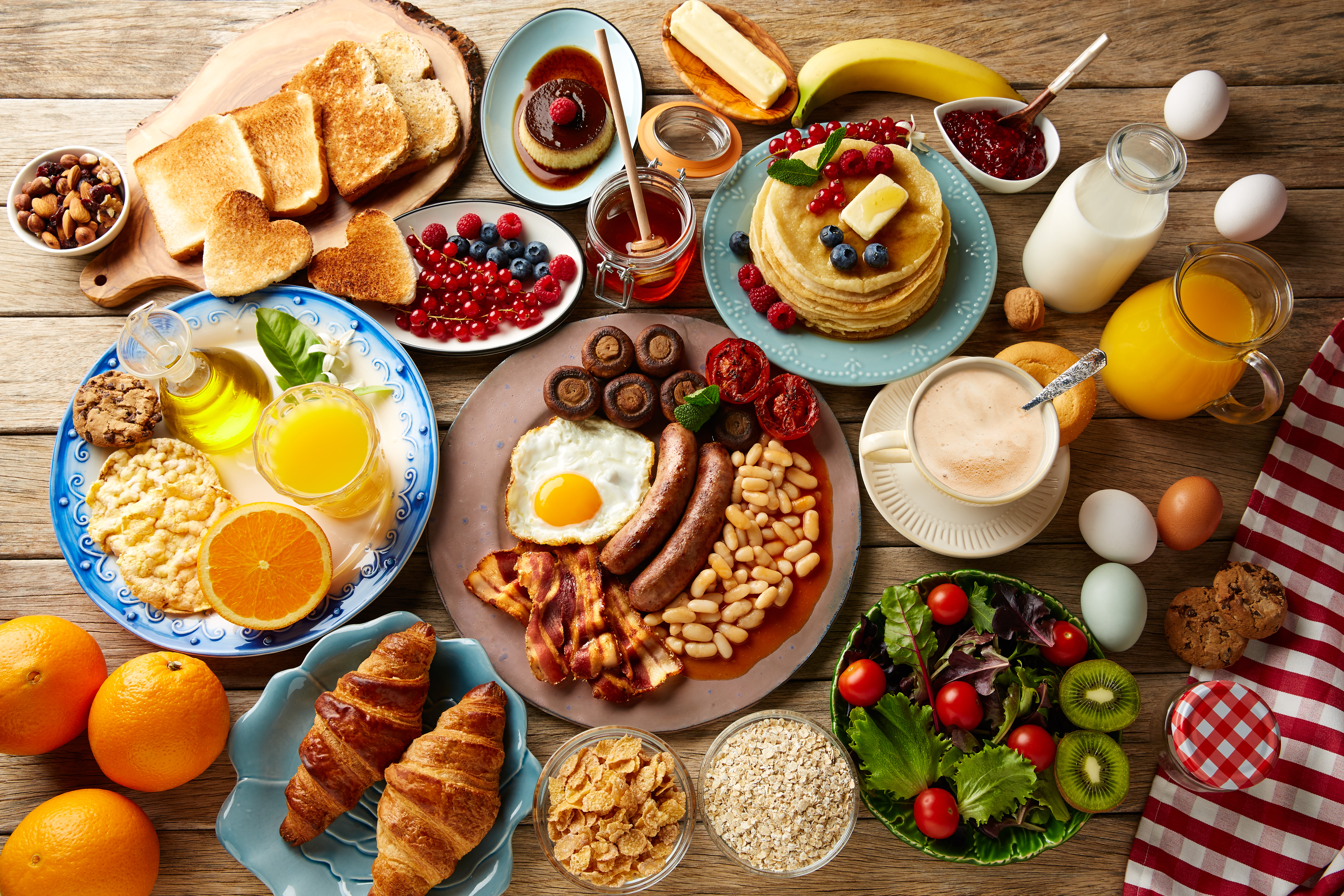 Several plates of breakfast food, including eggs, sausage, pancakes, toast, croissants, and fresh oranges seen from above.