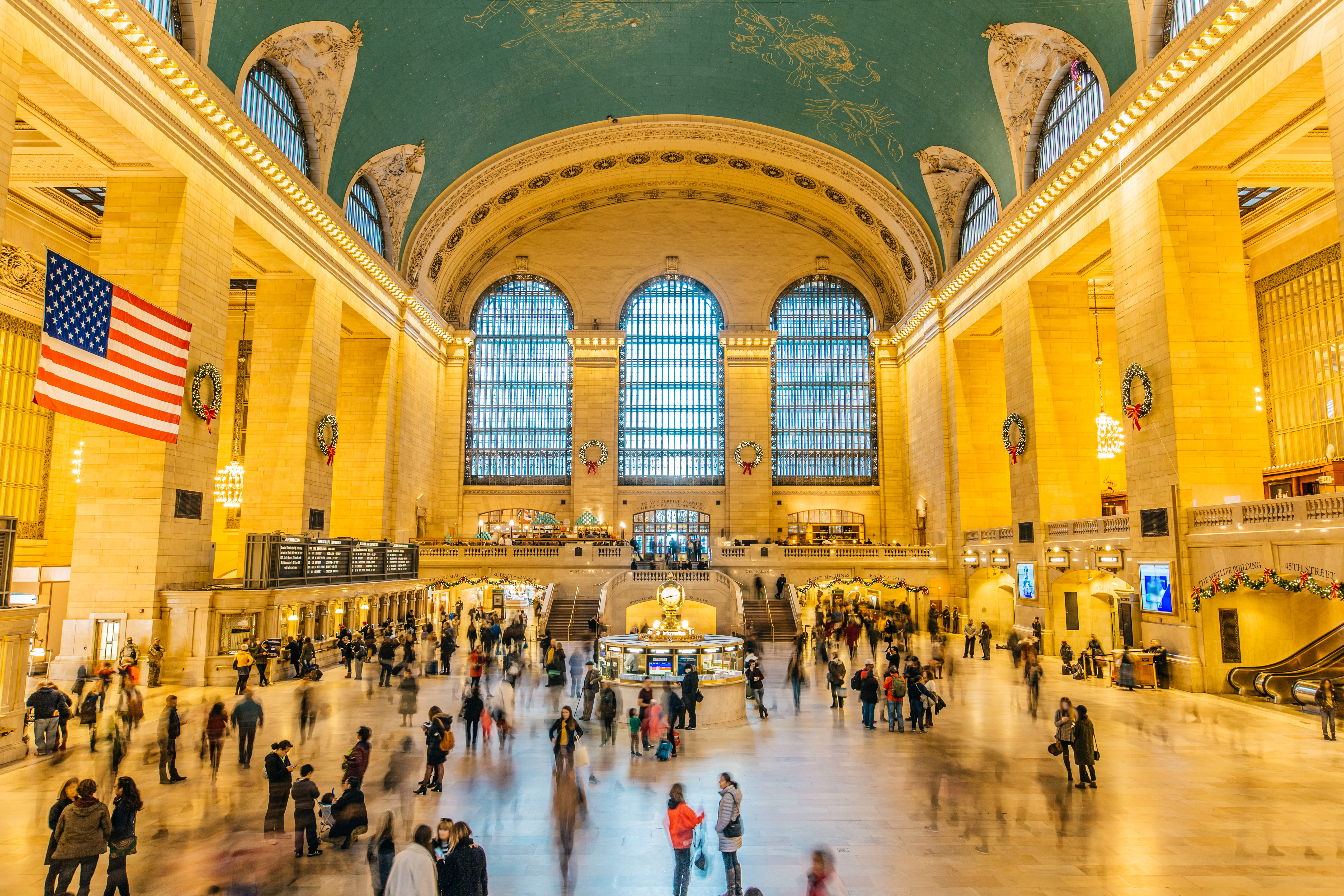 The main grand central terminal with a golden vaulted ceiling with green paint. Some people are seen walking inside the terminal.