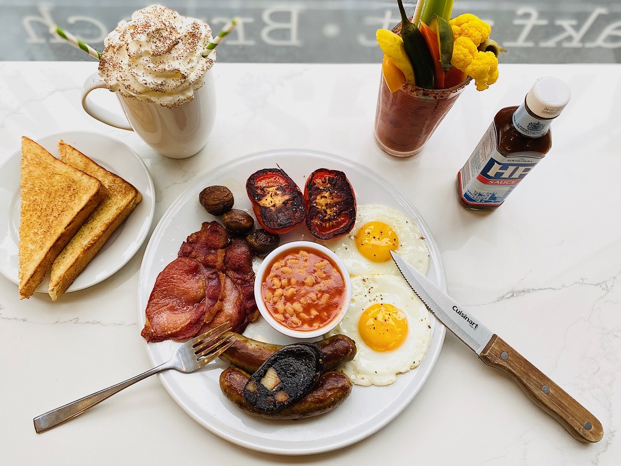 A white plate is packed with a container of beans, two fried eggs, two broiled tomatoes, rashers, black pudding and more, with a side of toast. This is the Full English at Kingsland Kitchen.