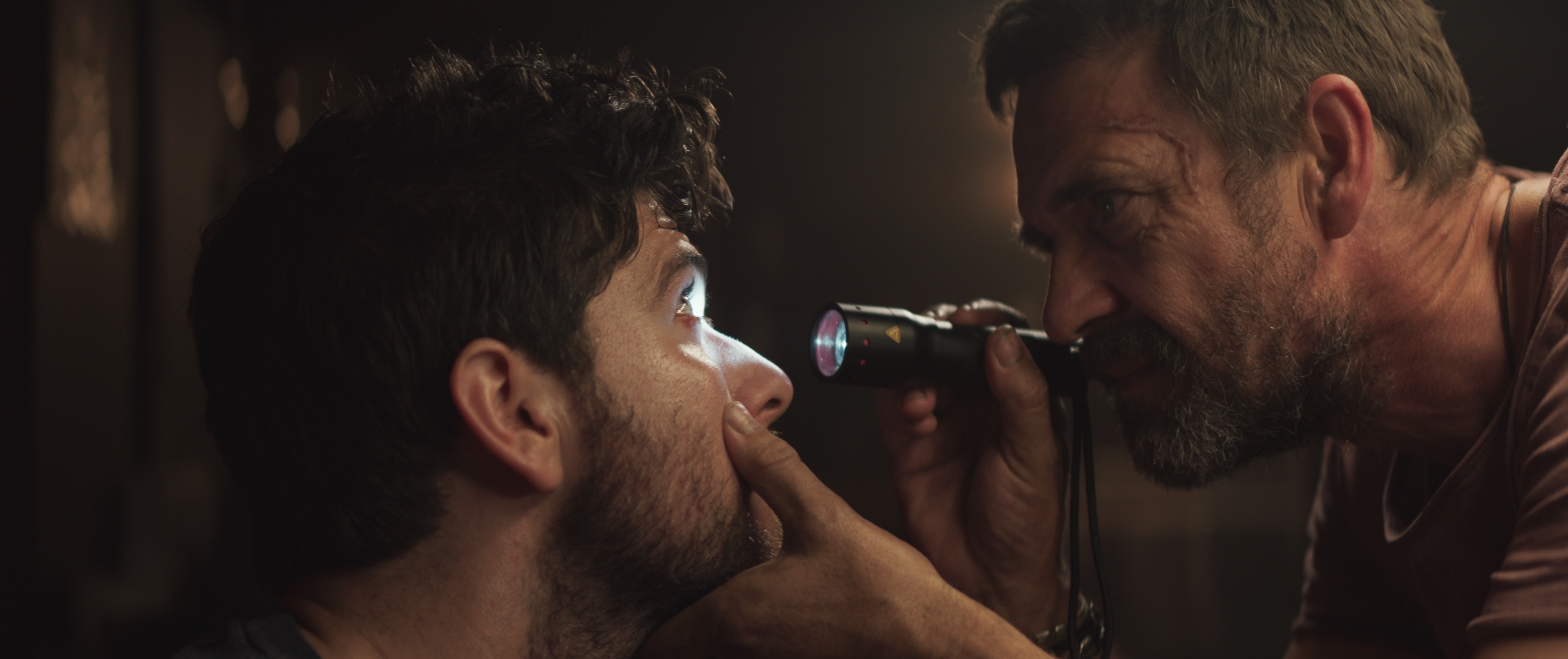 One grim looking-man shines a flashlight into another’s eyes while examining him for parasitic contagion in the horror movie Sea Fever.