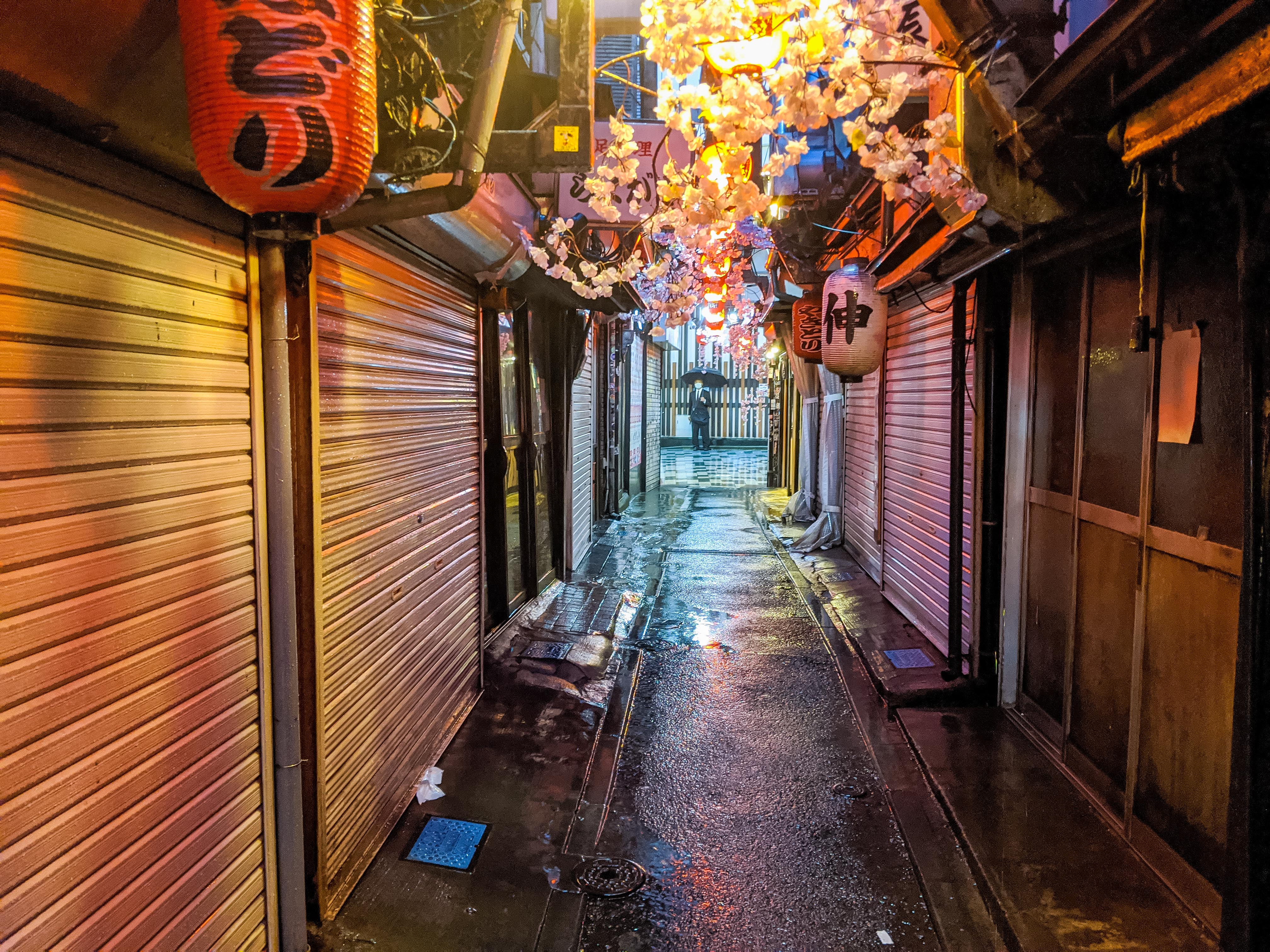 A slim alleyway, lit by hanging paper lanterns, where restaurants are closed behind steel shutters