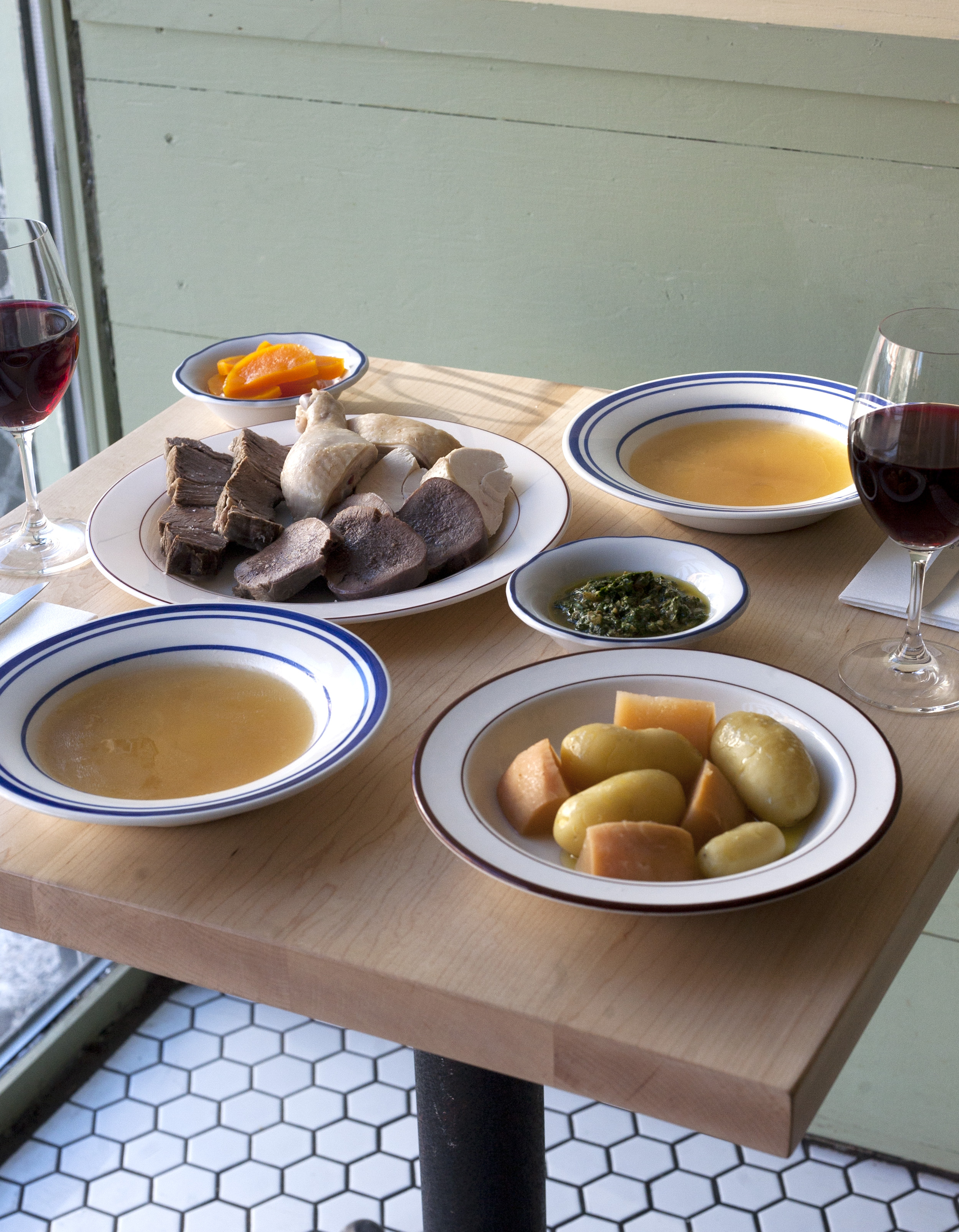 wood table with two glasses of wine, two bowls of soup and other plates with potatoes and meat