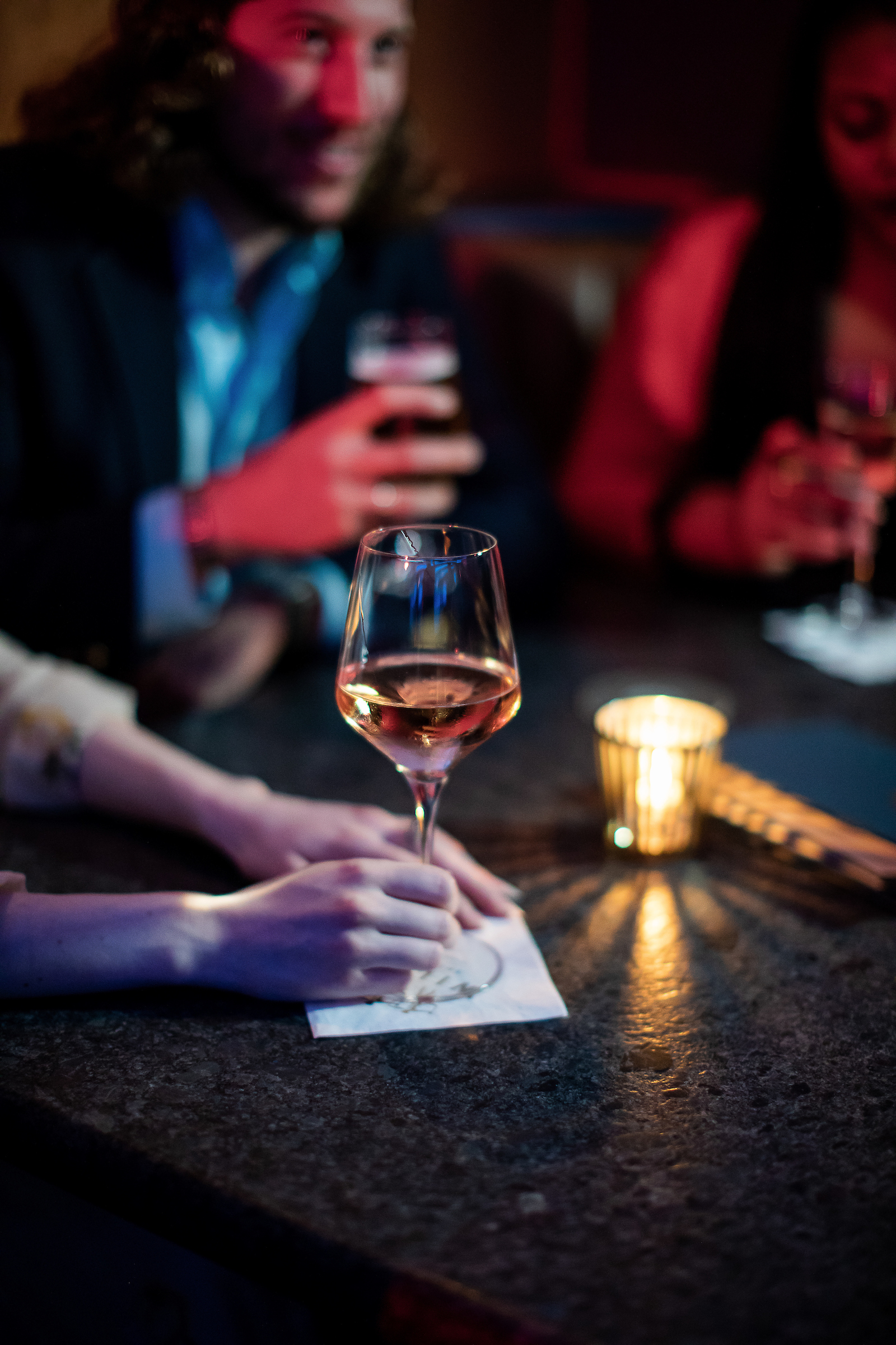 A woman’s hand holds the stem of a glass of wine on a table in a dark bar. 
