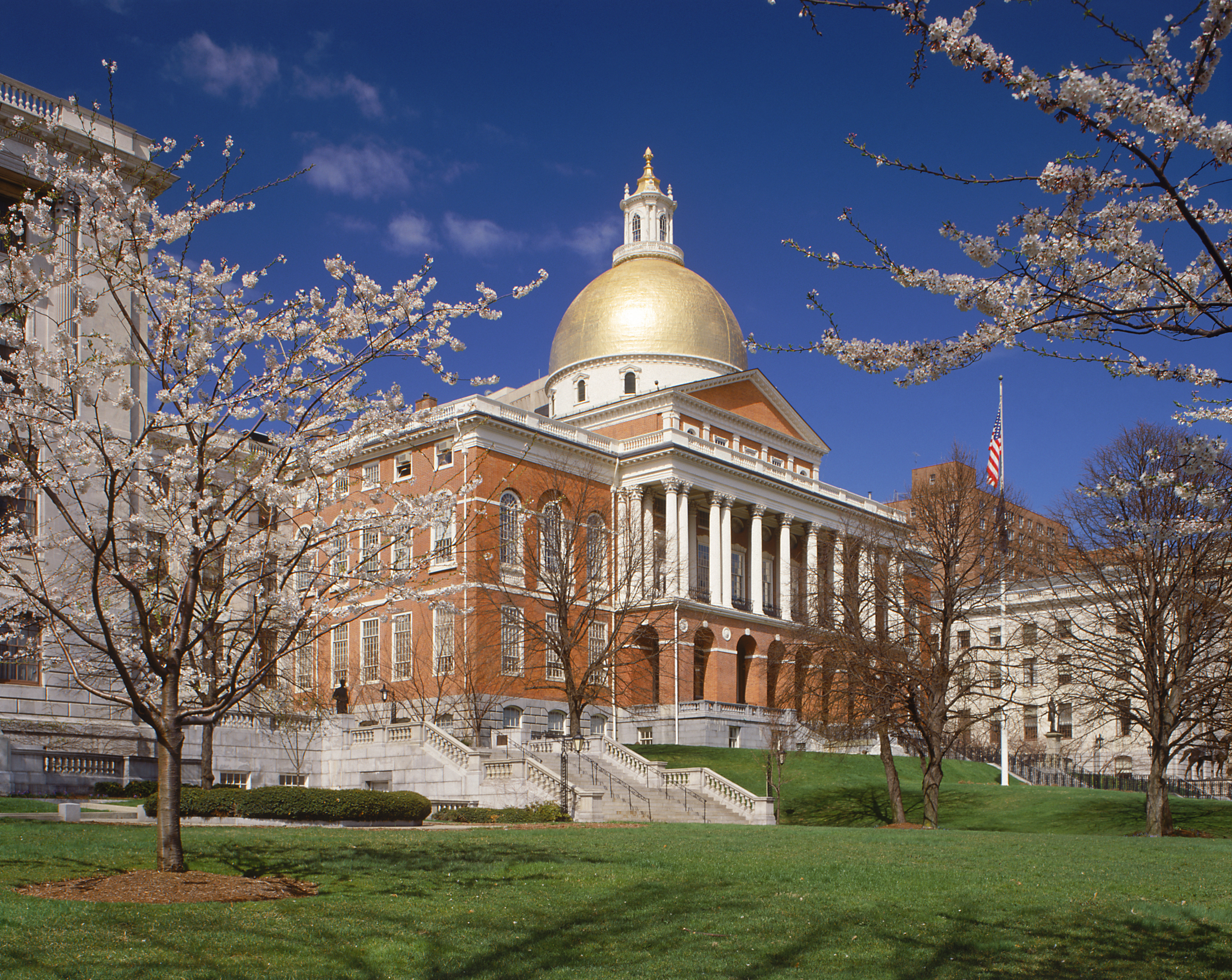 The exterior of the Massachusetts State House. The building is red brick and there is a gold dome. There are white columns on the facade.
