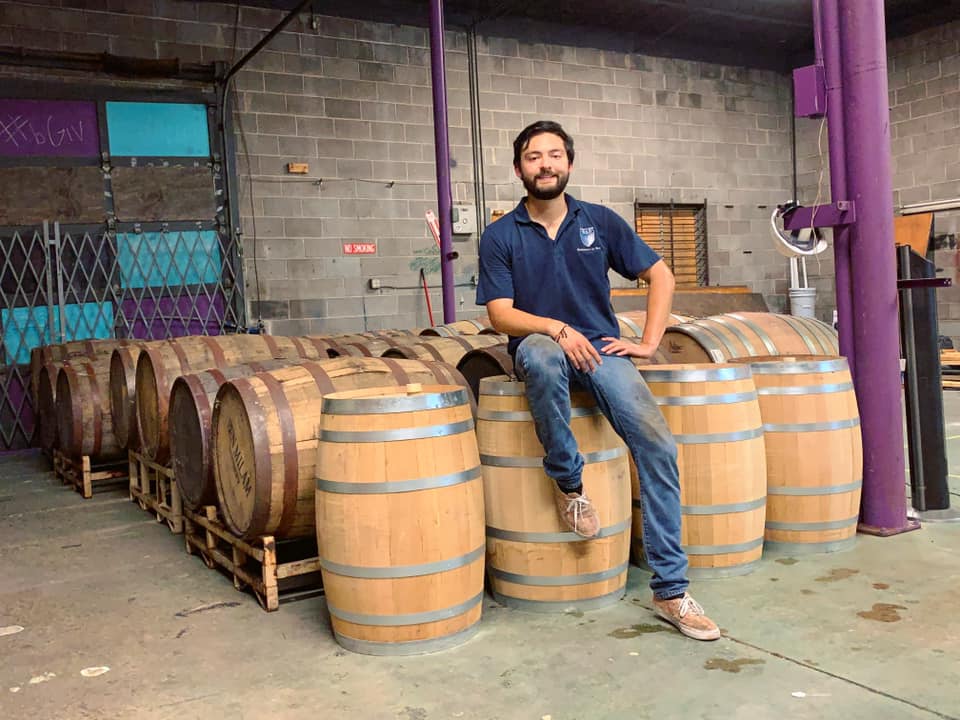 A man sitting on barrels of wine with with concrete bricks behind him that have been painted teal and purple