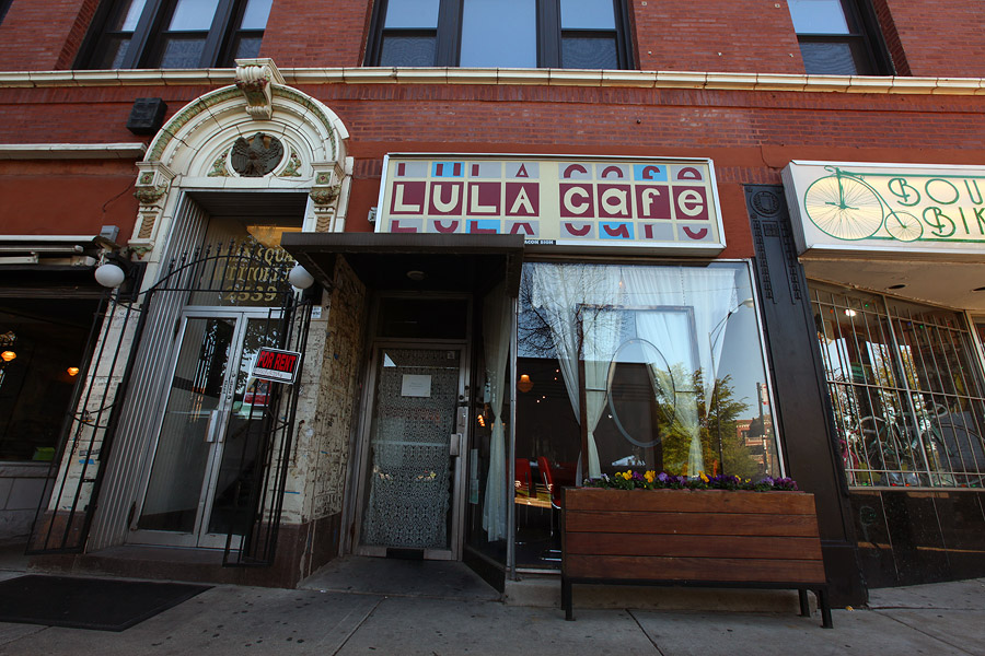 A storefront inside a brick building from the street. White curtains are visible through a large window and a sign overhead reads “Lula Cafe.”
