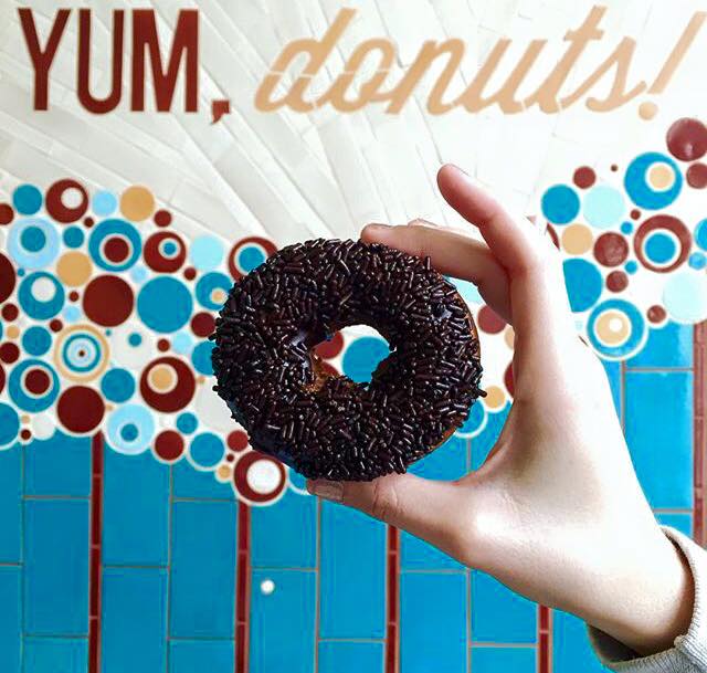 A hand holds a chocolate frosting and sprinkle topped doughnut in front of a sign that says, “Yum, donuts!”