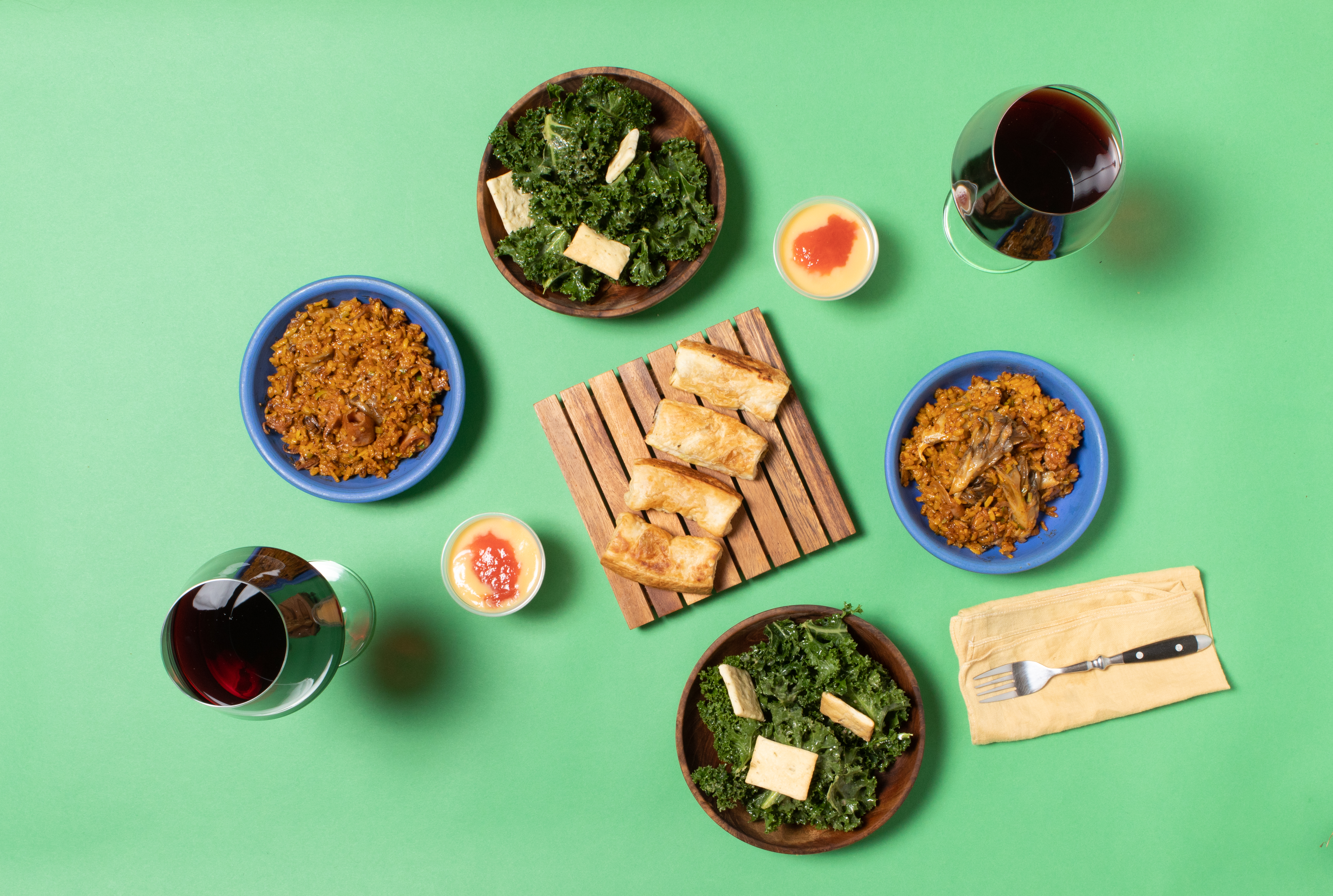 Top-down view of bowls of paella, glasses of red wine, and kale salad against a green background