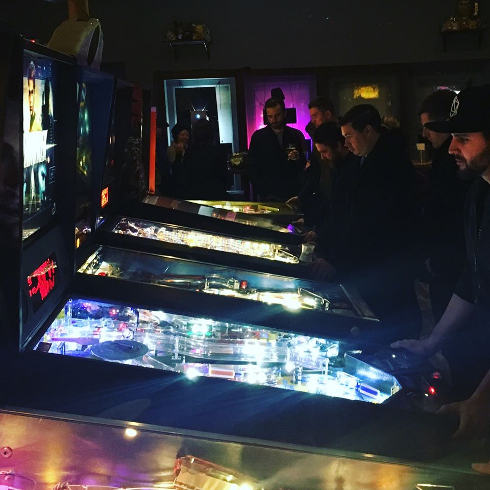 In a dark room, a line of lit up pinball machines with players