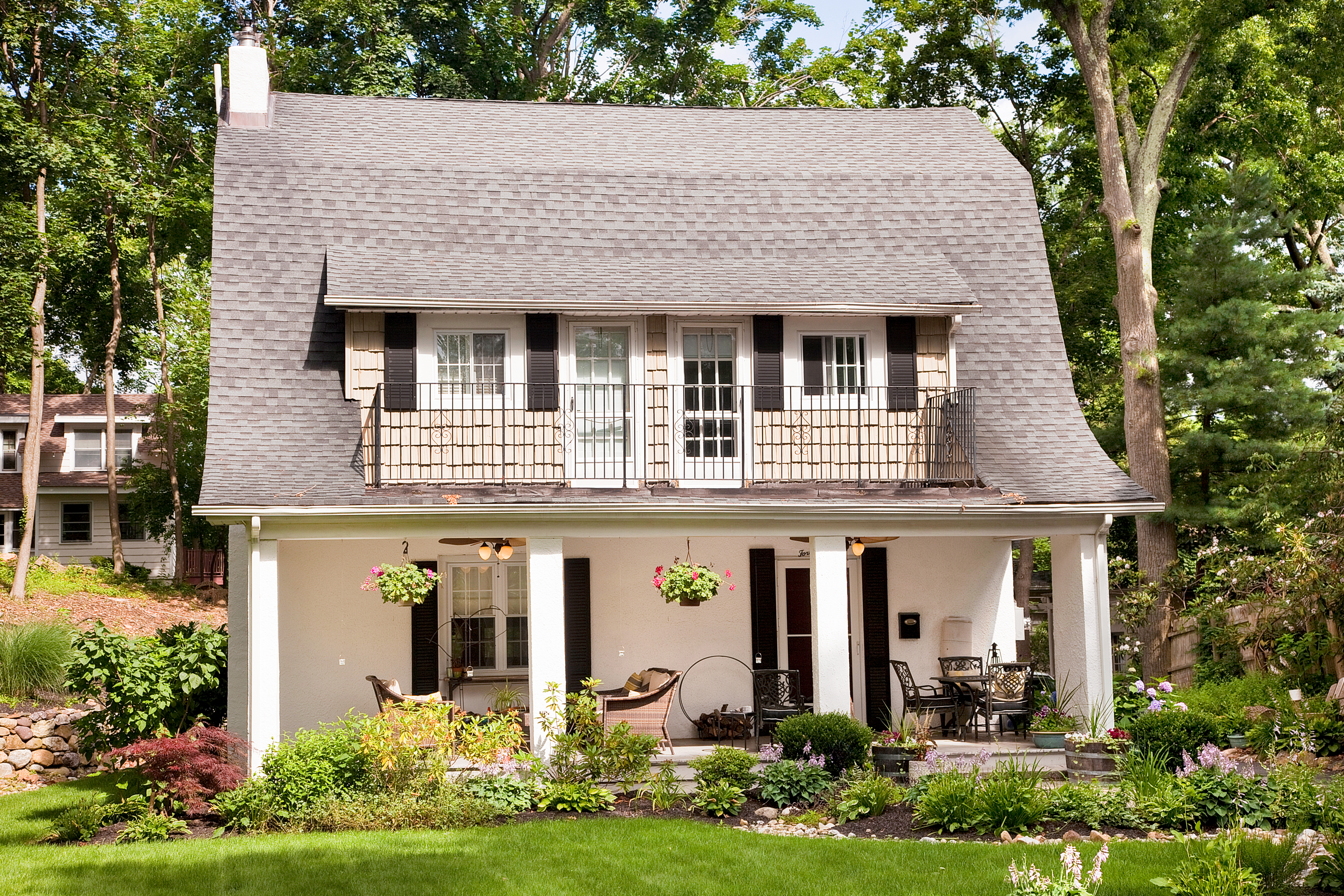 A two-story white and tan cottage with black window finishings, a front porch with furniture, a chimney, and a well-manicured lawn and foliage