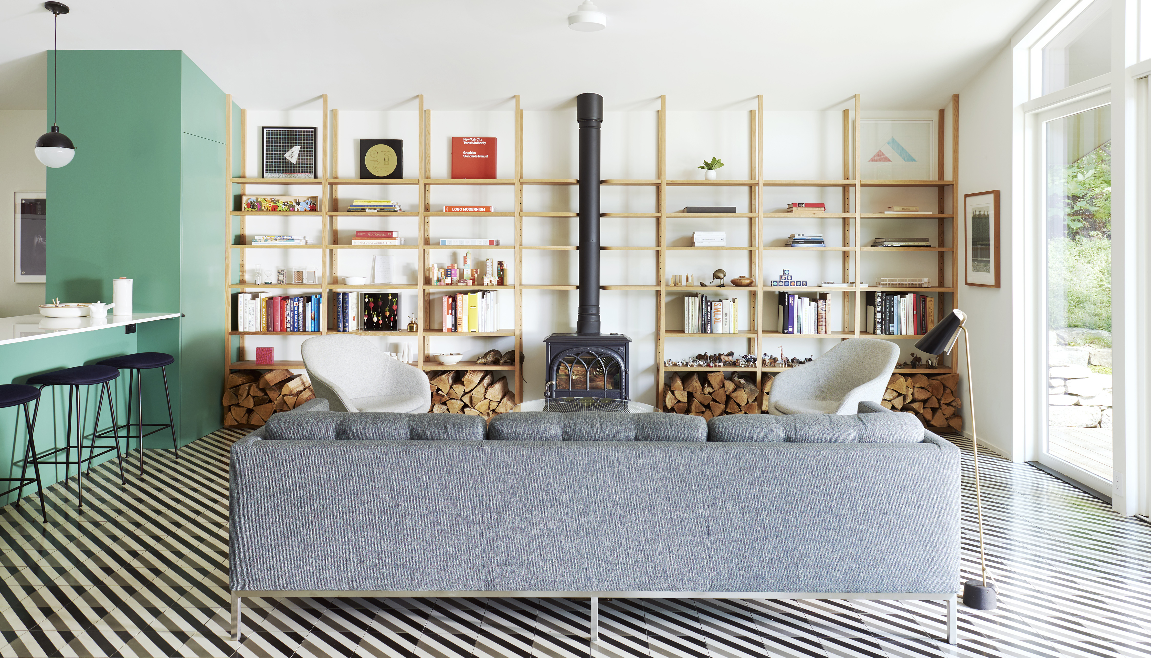 A grey couch sits in front of a living room with a wood burning stove as the focal point. To the stoves left and right are floor-to-ceiling wooden shelves that are minimally stacked with books, art, and colorful trinkets. The floor is a tiled diagonal white, grey and black striped pattern.