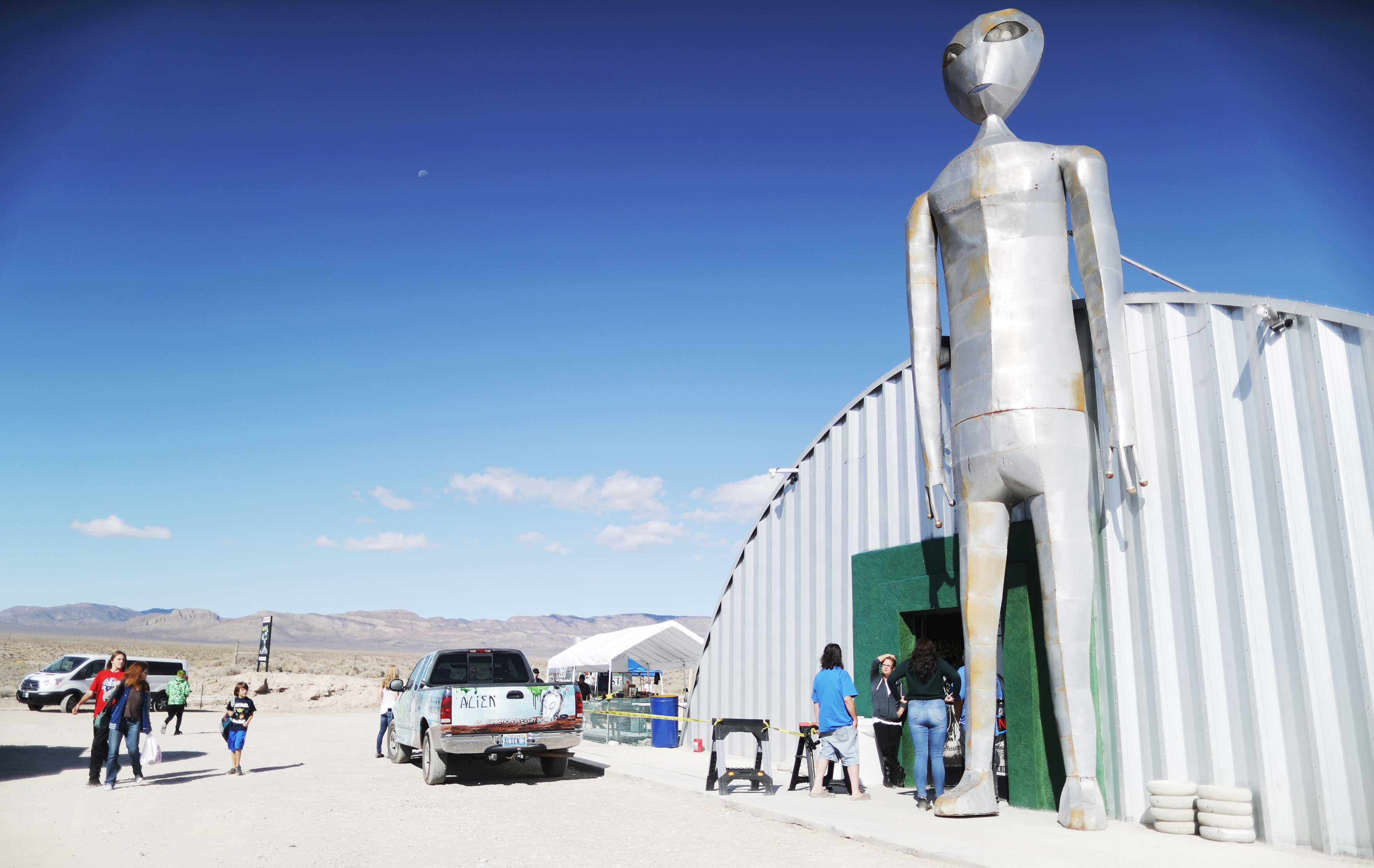 A giant metal sculpture of an alien stands outside a trailer in the desert.