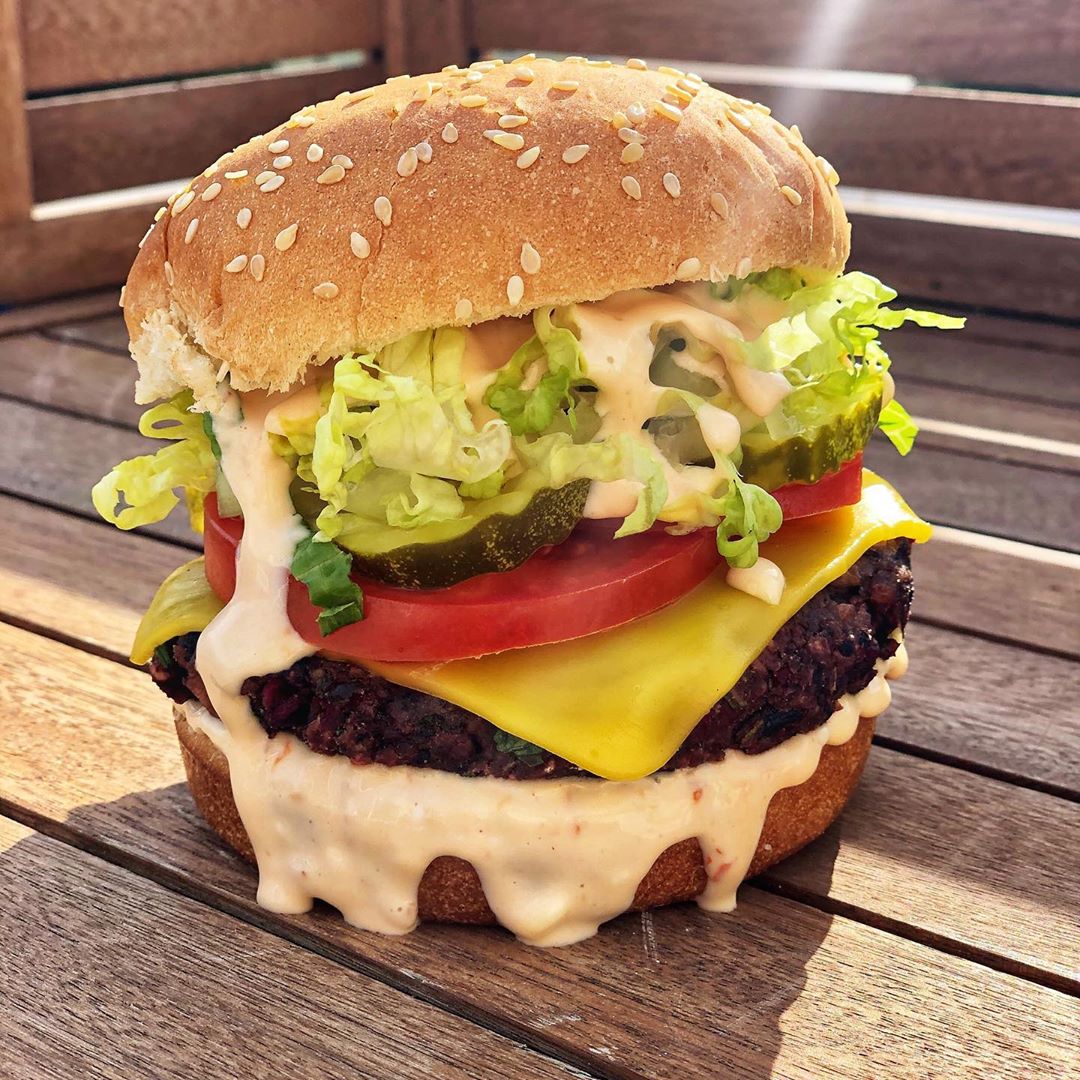 A vegan burger with tomato, lettuce, pickles, “cheese,” and a creamy sauce sits on what appears to be a picnic table
