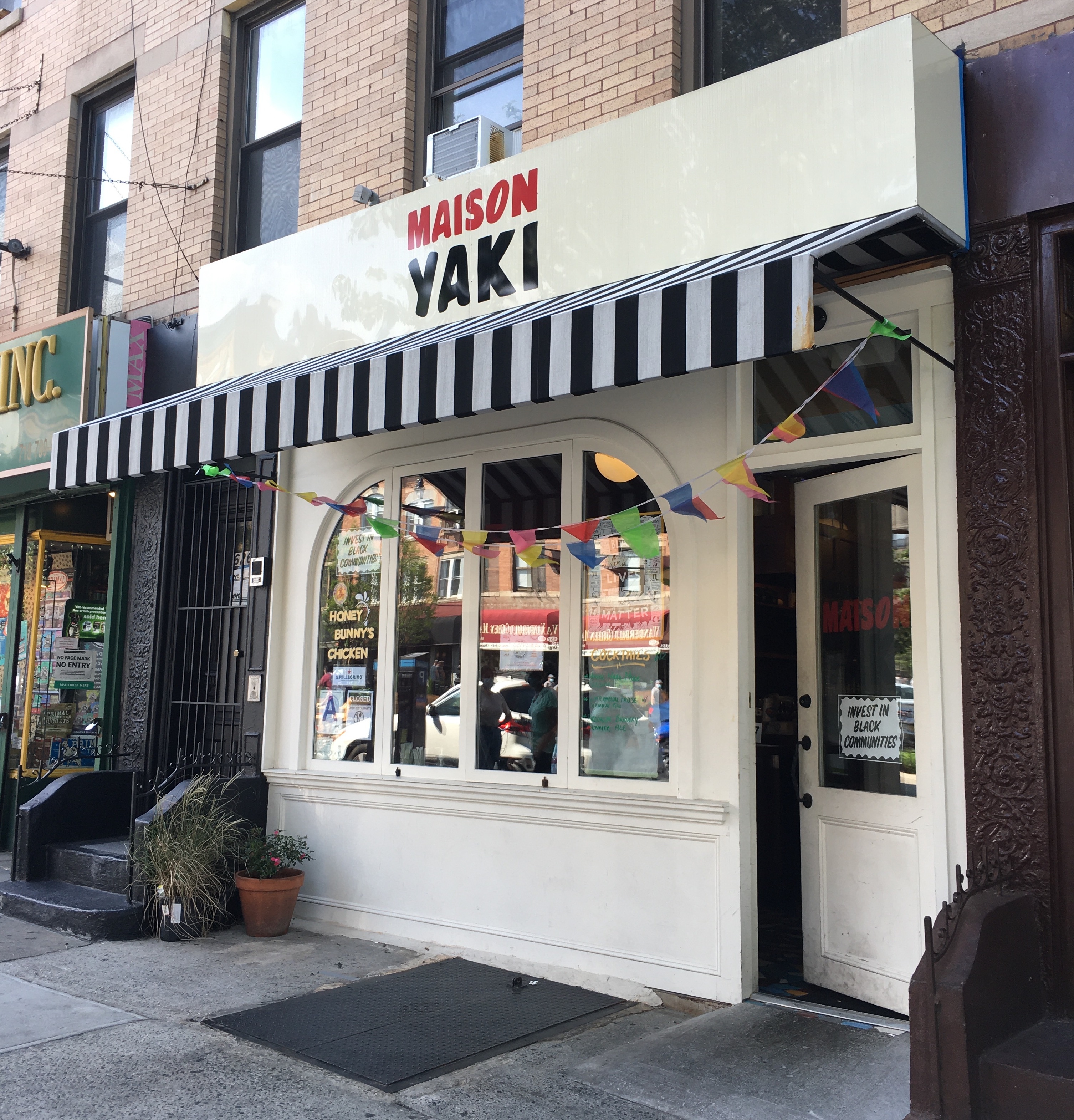 The front of a restaurant with a striped awning, colorful flags, and a sign that reads “Maison Yaki”