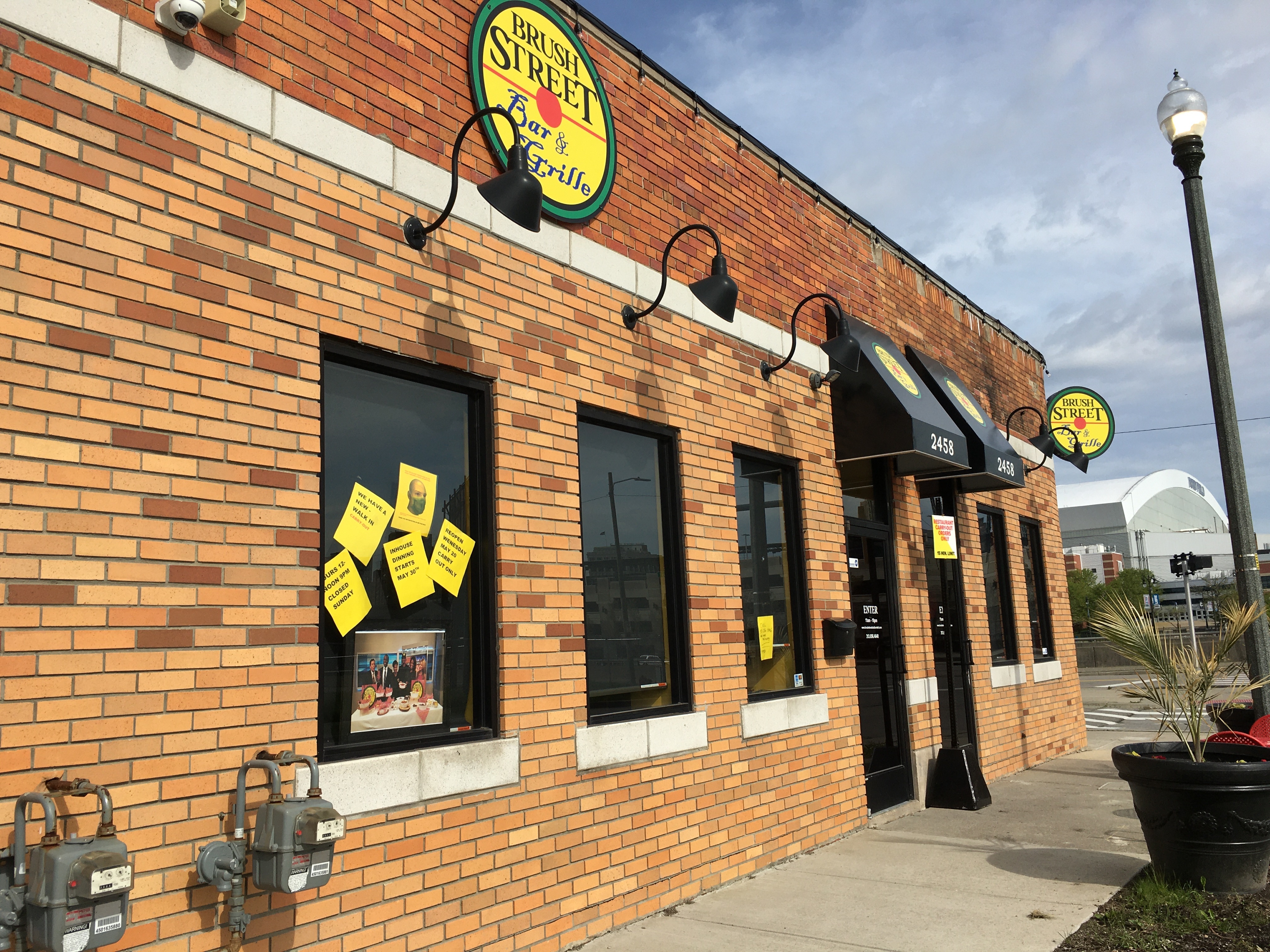 Brush Street Bar &amp; Grille is located in a brick building with a curved corner storefront. The restaurant is shown on a sunny day with yellow paper signs plastered in the windows related to coronavirus operations.
