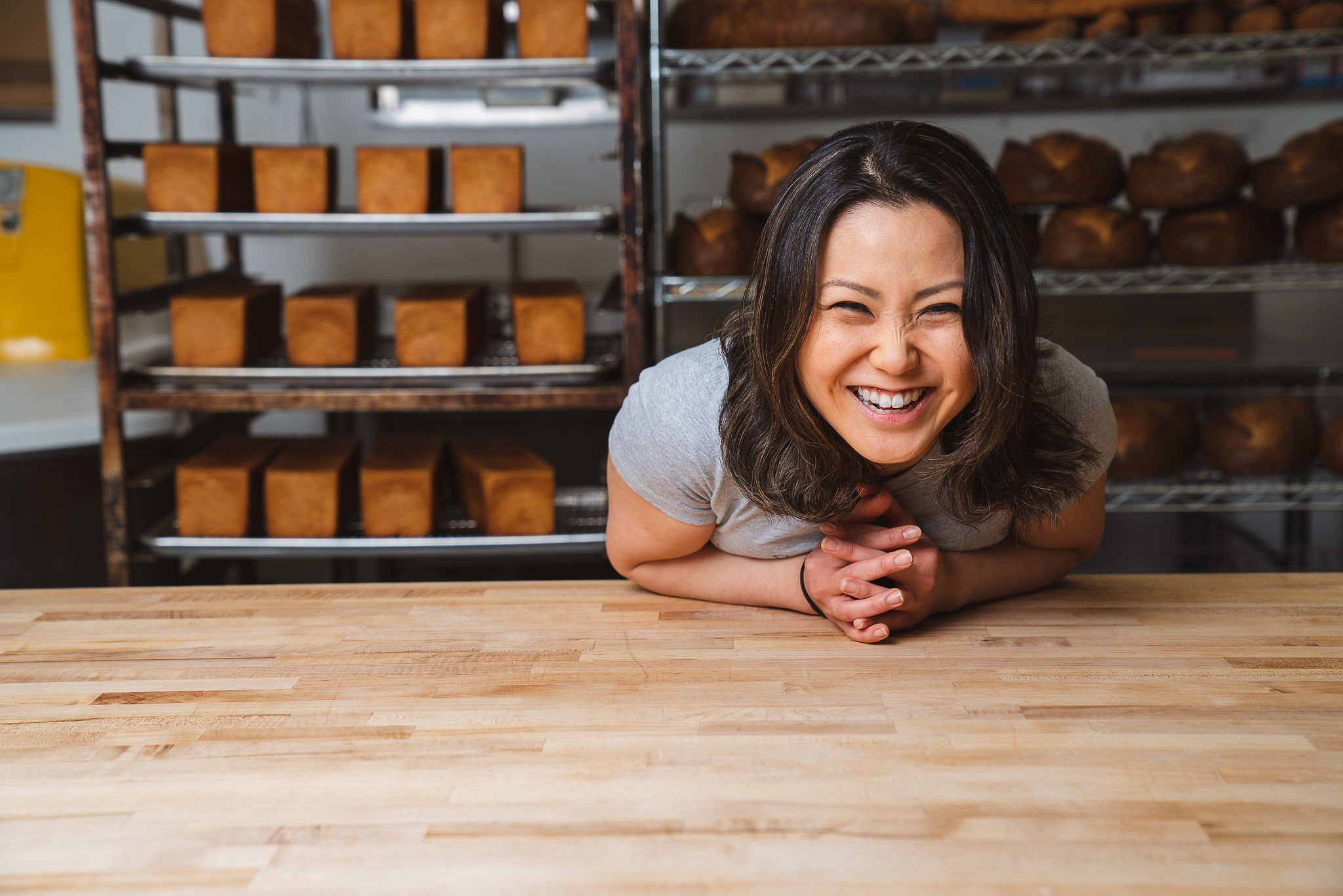 A woman with long and black hair smiles and leans over a wooden table with loaves of bread on racks in the background.
