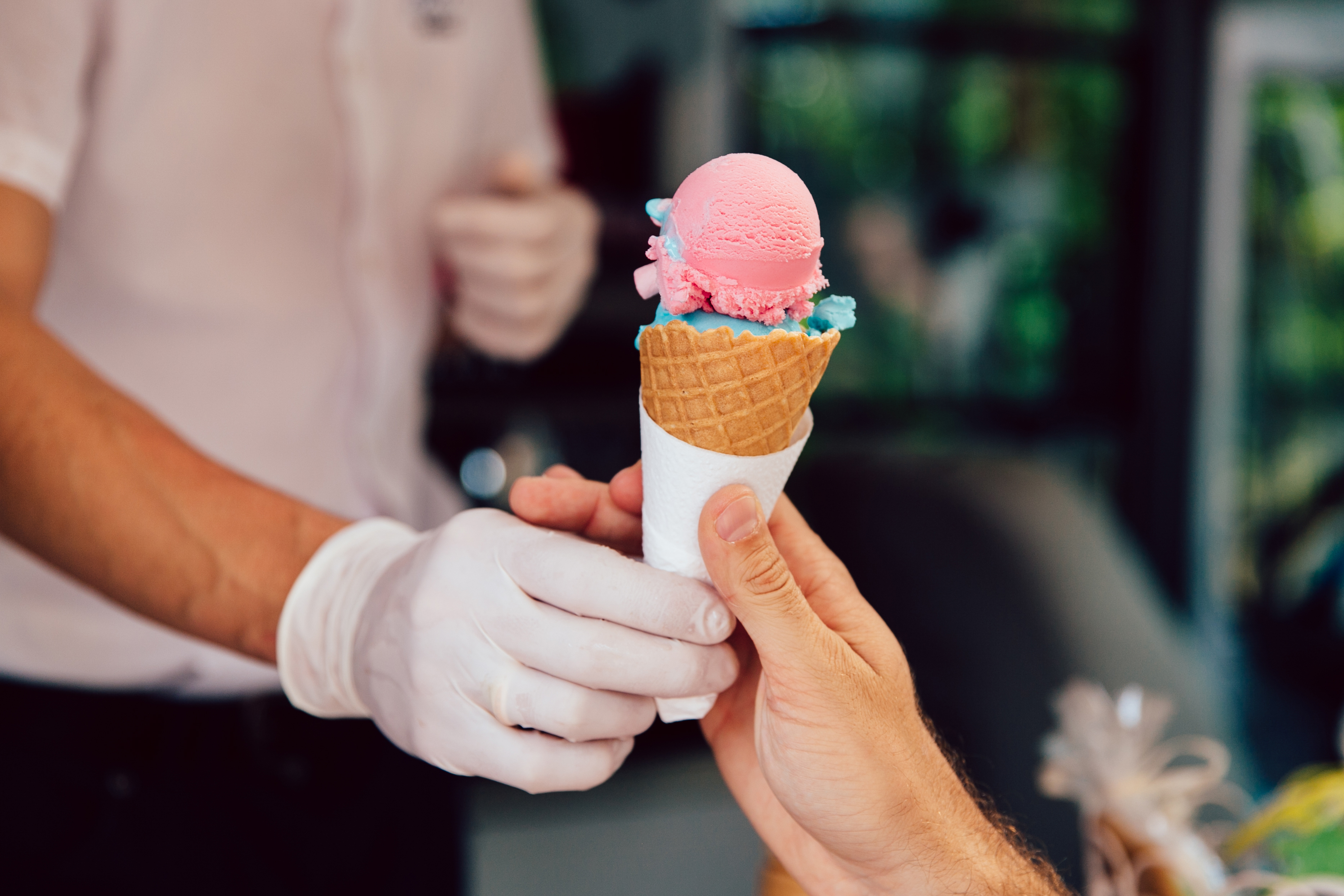 A gloved hand passes off an ice cream cone to an ungloved hand. It’s filled with pink and blue scoops of ice cream. 